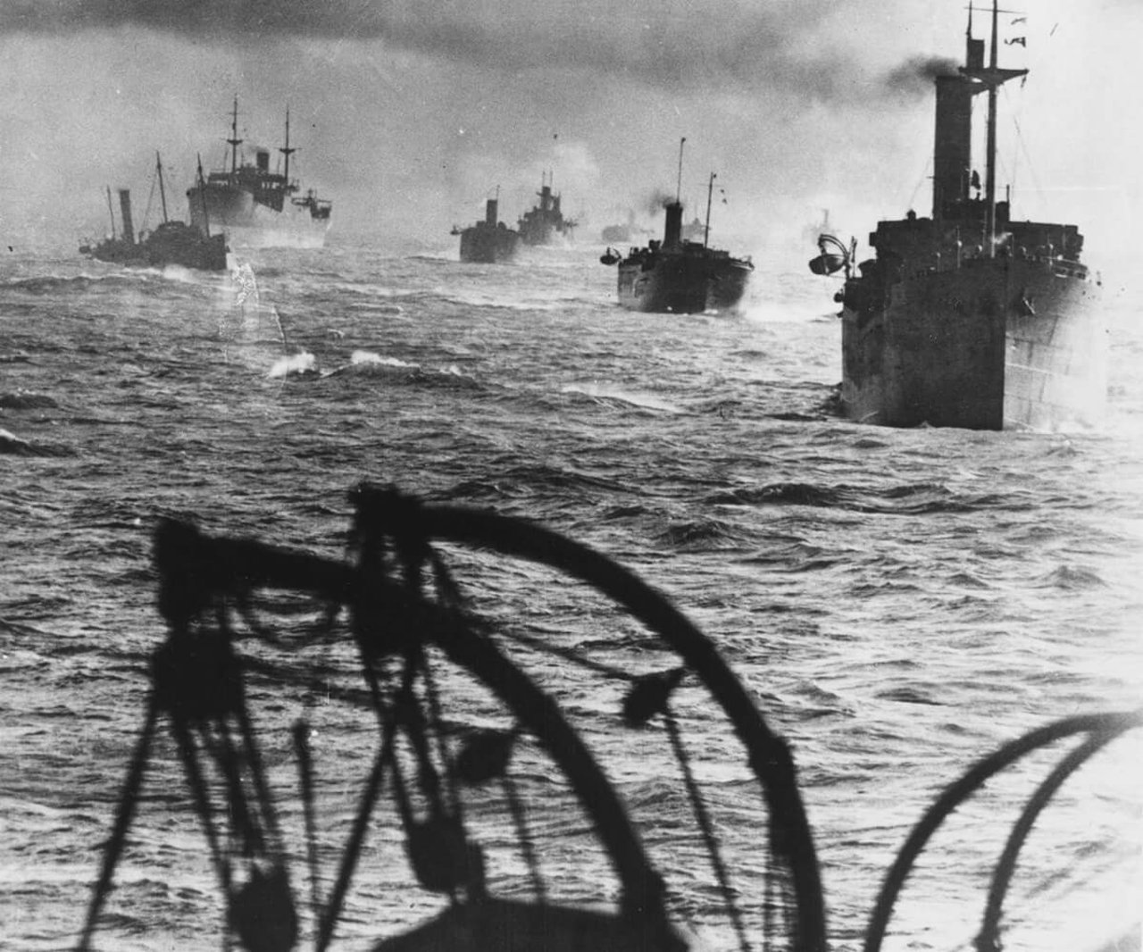 Black and white photo taking from a ship showing many war ships travelling in one direction.