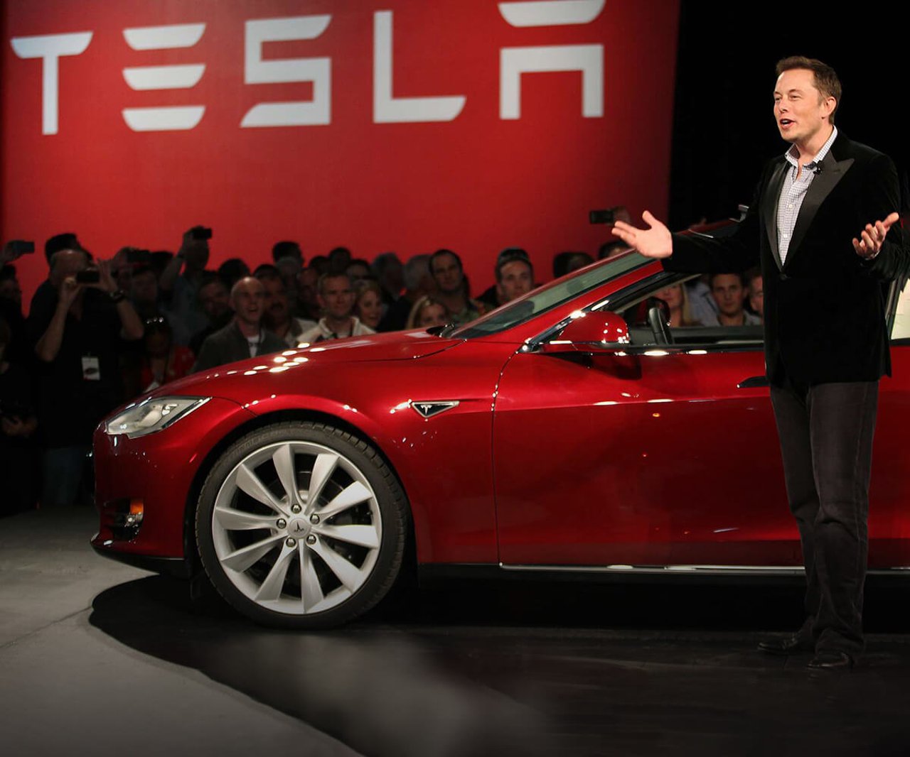 Elon Musk delivering a speech stood in front on a red Tesla with the Tesla logo in the background.
