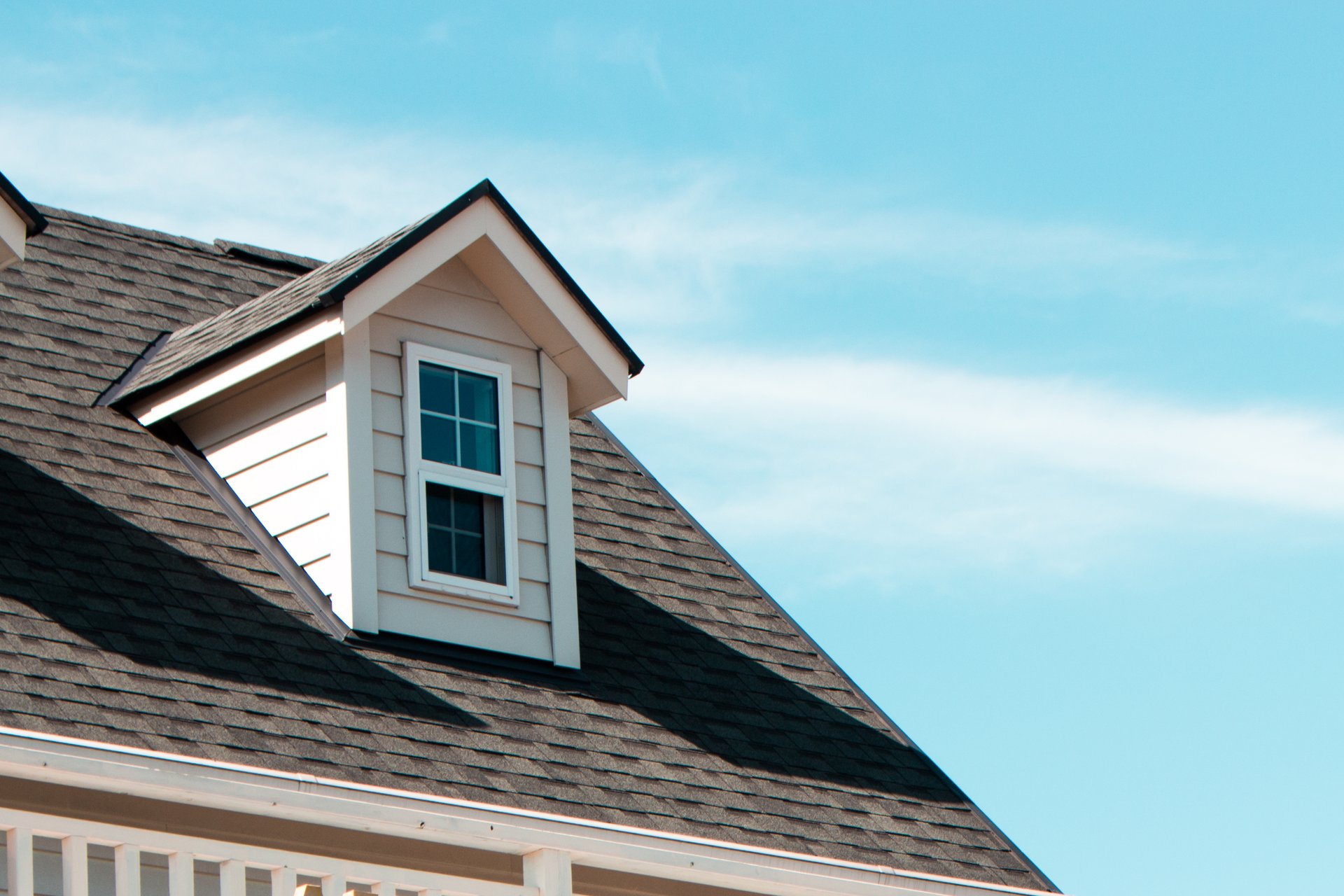 alt="Close up of a house roof and roof window in blue sky with clouds"