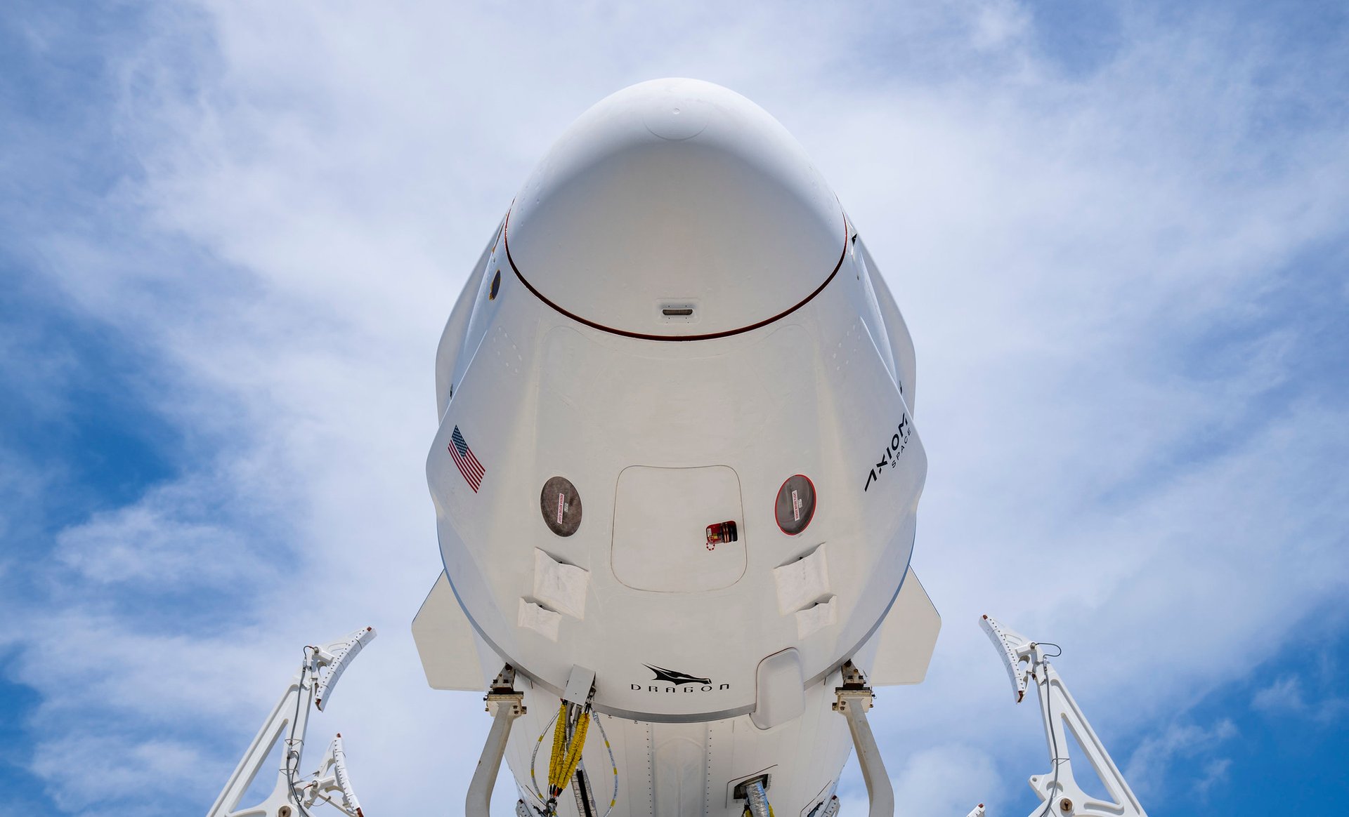 Close-up view from below of a SpaceX Dragon spacecraft against a blue, cloudy sky, showing its rounded nose, markings, and support structures.