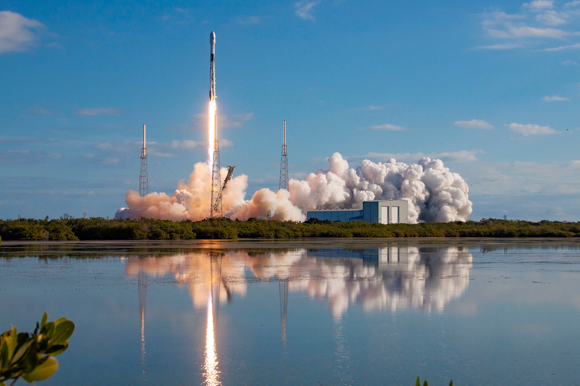 A SpaceX Falcon 9 rocket launches a Starlink mission from a coastal launchpad, with bright flames, thick smoke, and reflections visible in the water below.