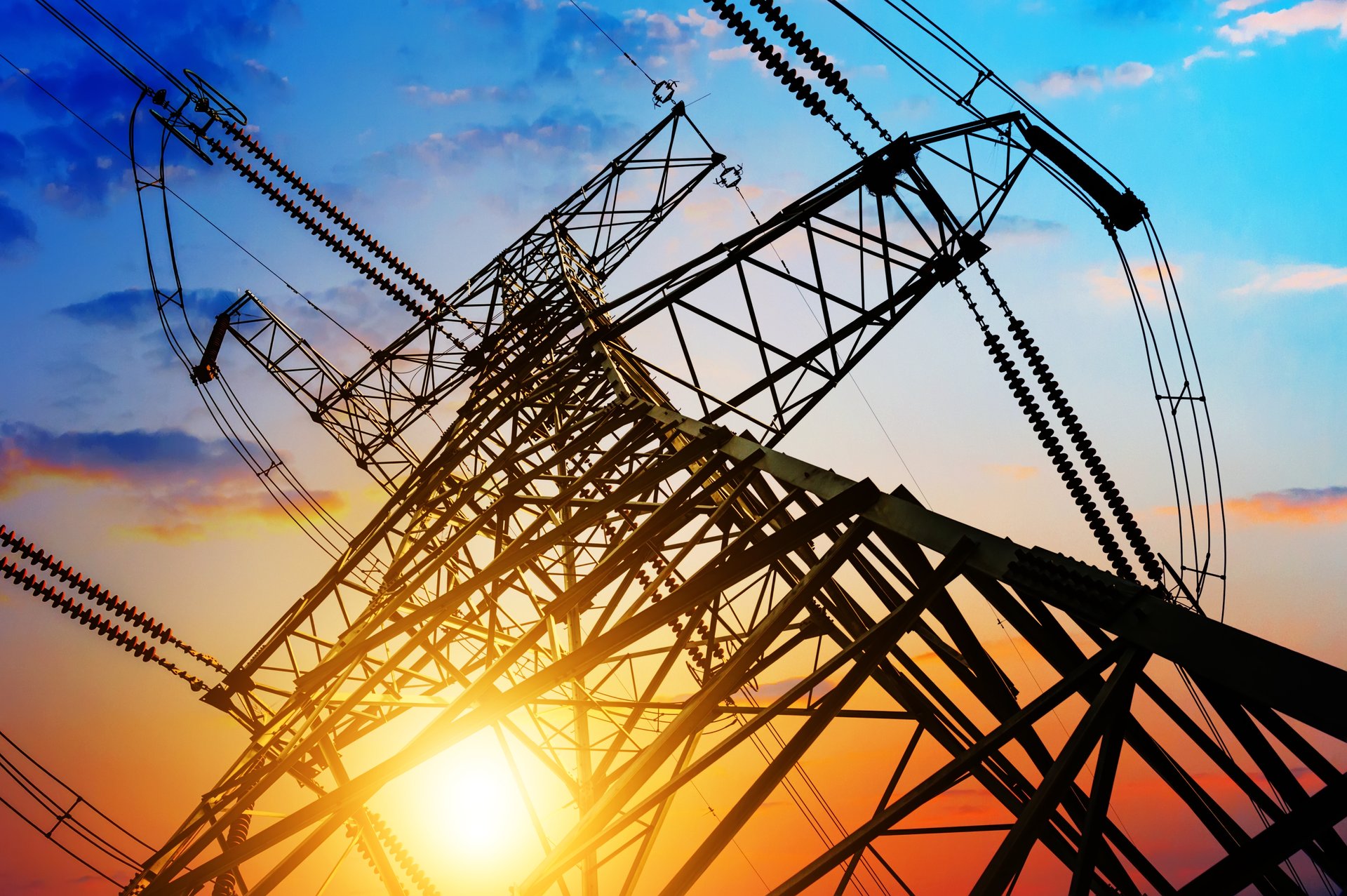 Low-angle view of a high-voltage electricity transmission tower silhouetted against a colourful sunset sky, with power lines and insulators stretching outward in multiple directions.