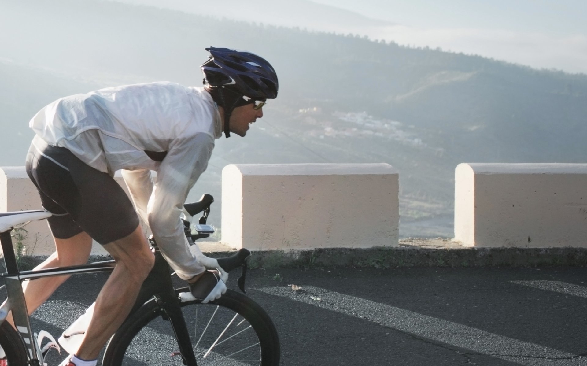Photograph of a cyclist wearing a helmet and white jacket riding a road bike uphill on a mountain road with concrete barriers. Background shows misty hills and valleys, highlighting a challenging outdoor cycling activity.