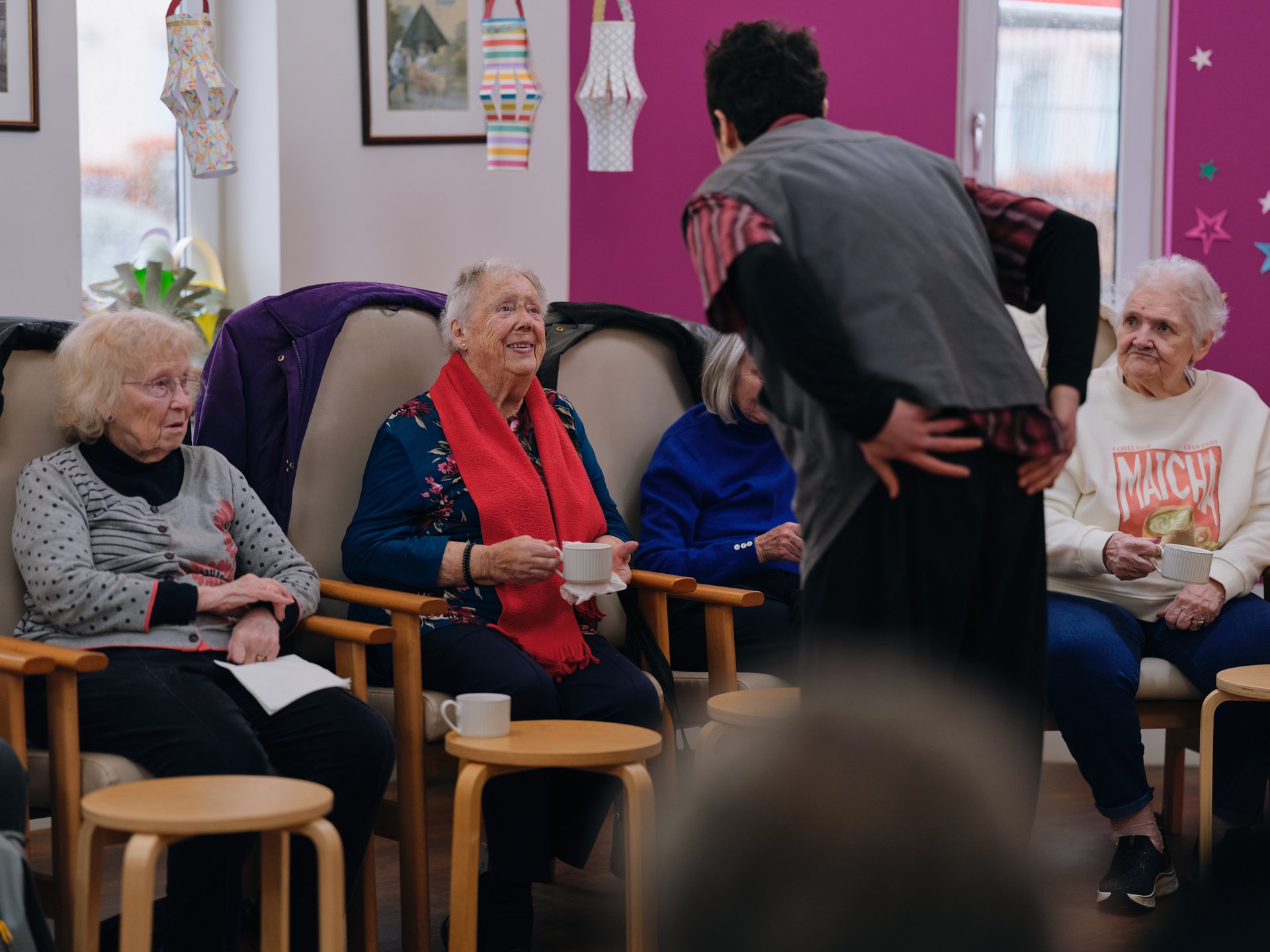 Photograph of a group of elderly women seated in a circle in a brightly coloured room, engaging in conversation with a standing woman facilitating the discussion.