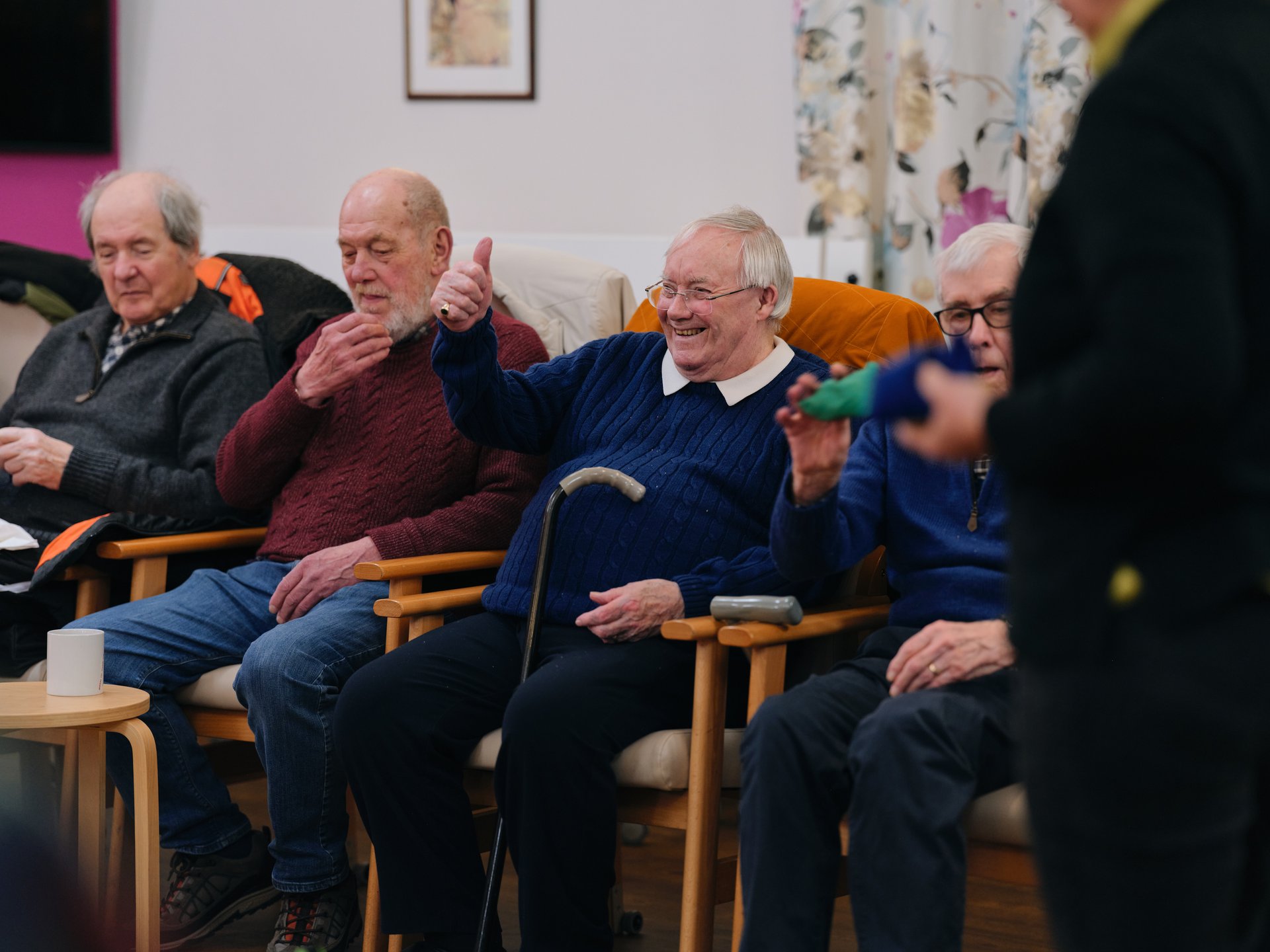 Elderly men seated in a row of armchairs in a communal room, engaging in conversation and gesturing with hands.