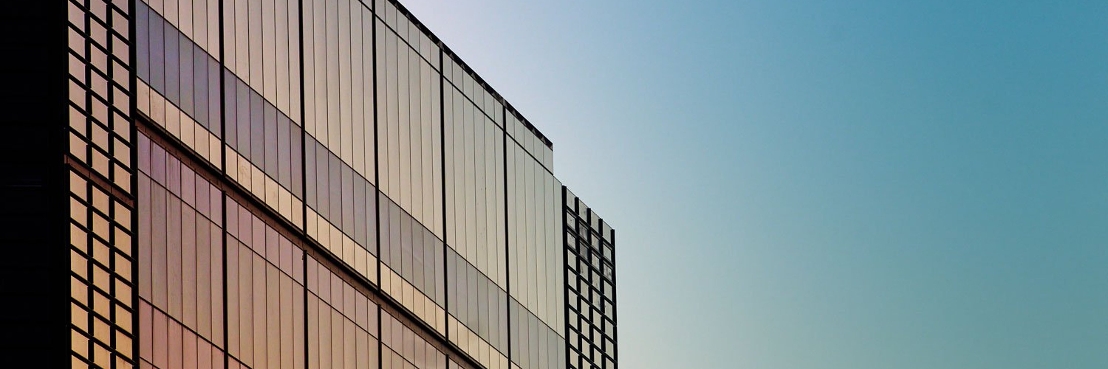 The side of a glass office building at with sunset colours reflecting on the glass against a clear blue sky.