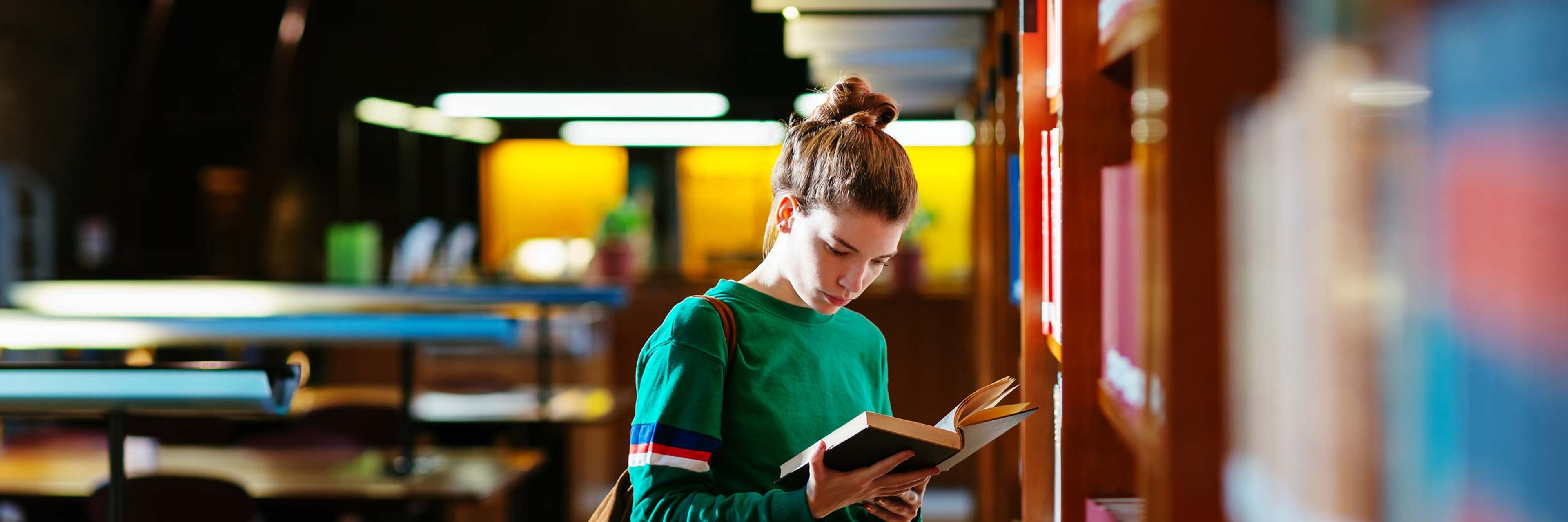 A young woman standing reading a book inside a library.