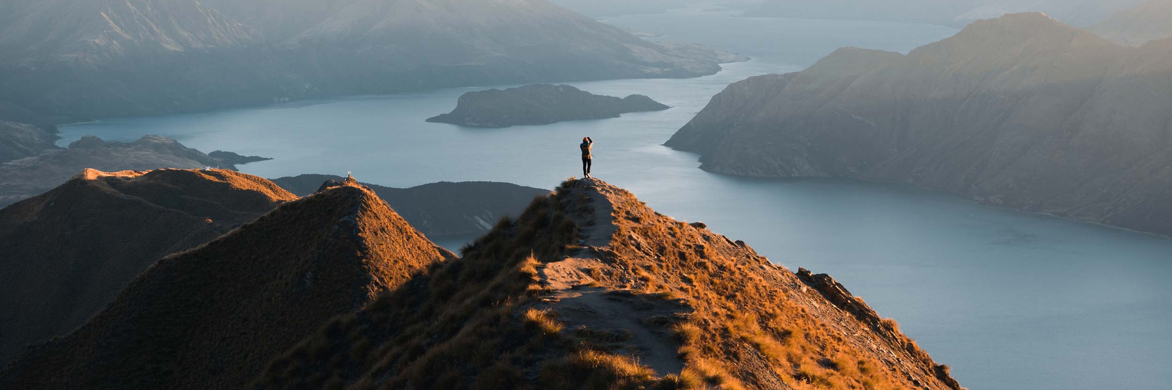 A silouette of a walker on top of a mountain looking over a lake in the valley below.