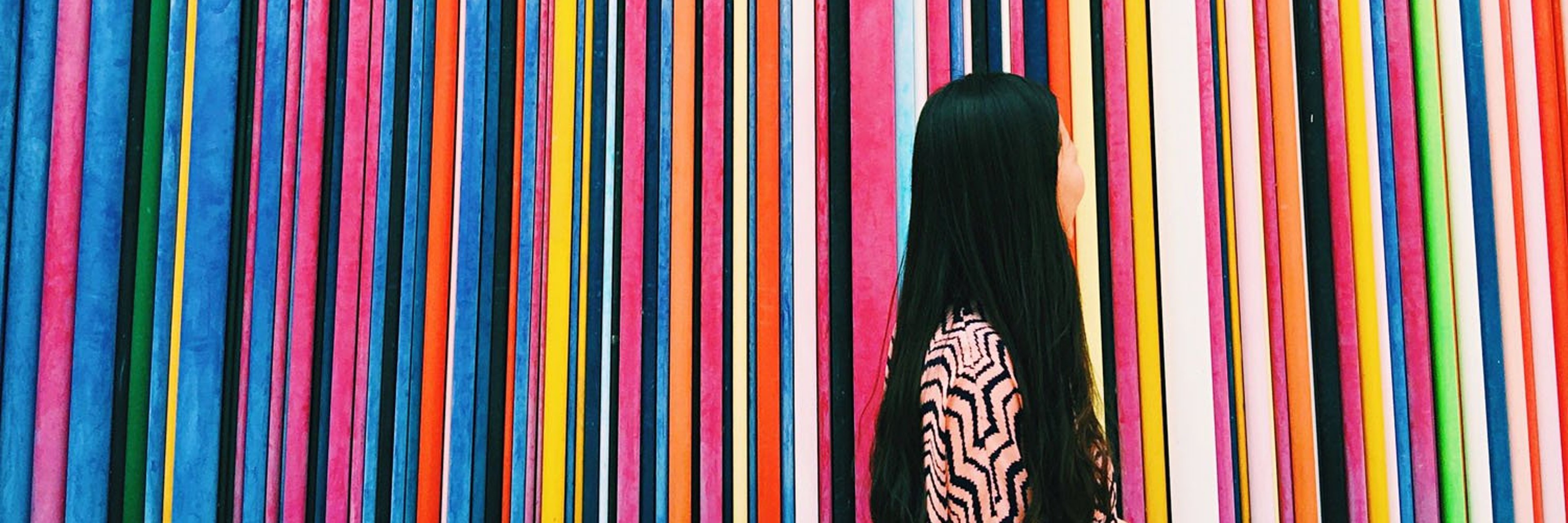 A woman with long black hair standing in front of a colorful fabric wall displaying multicolored lines.