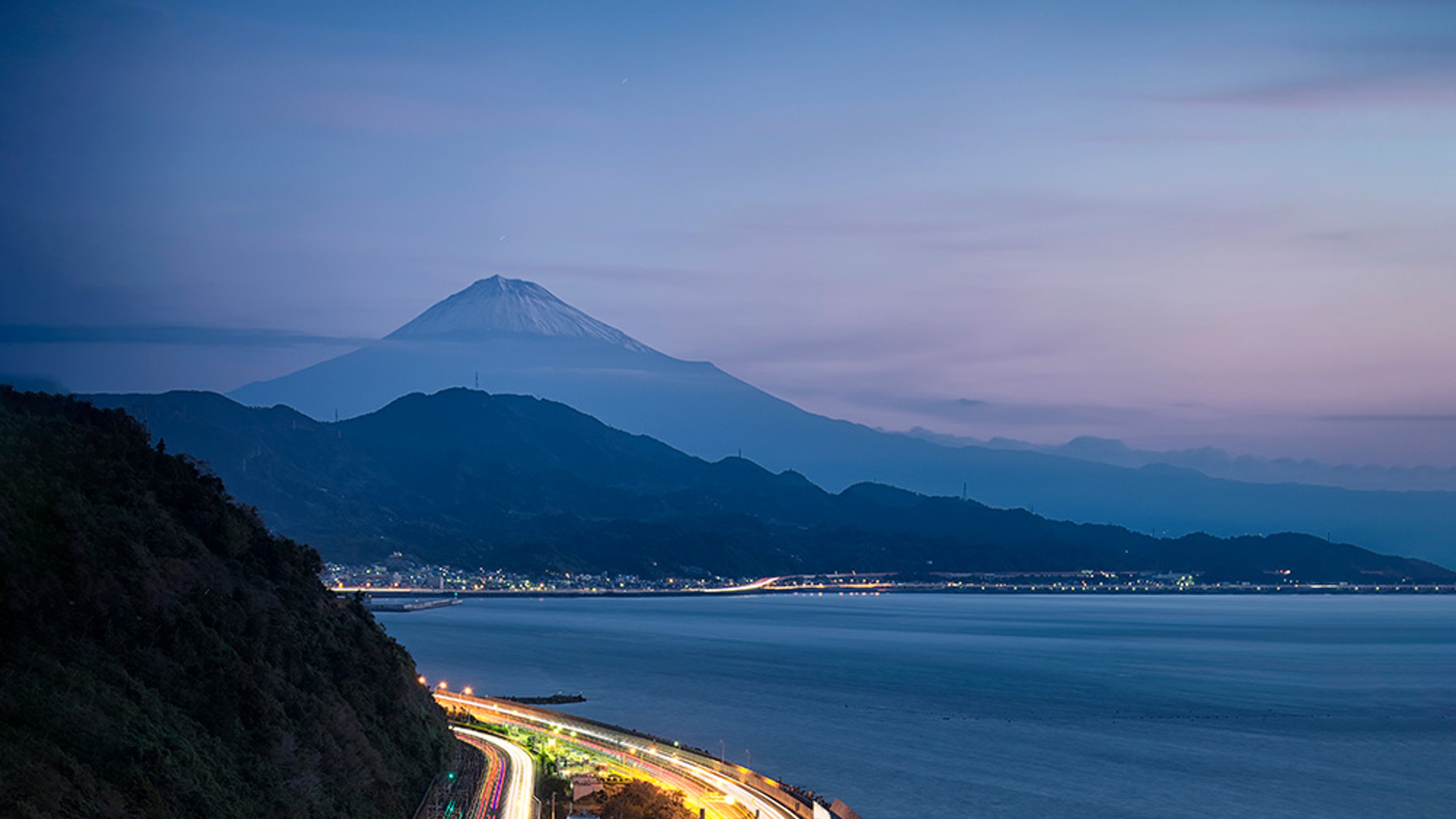 Satta Pass at dawn in Shizuoka, Japan. The winding coastal road with car light trails,  and Mount Fuji in the background under a clear, early morning sky.