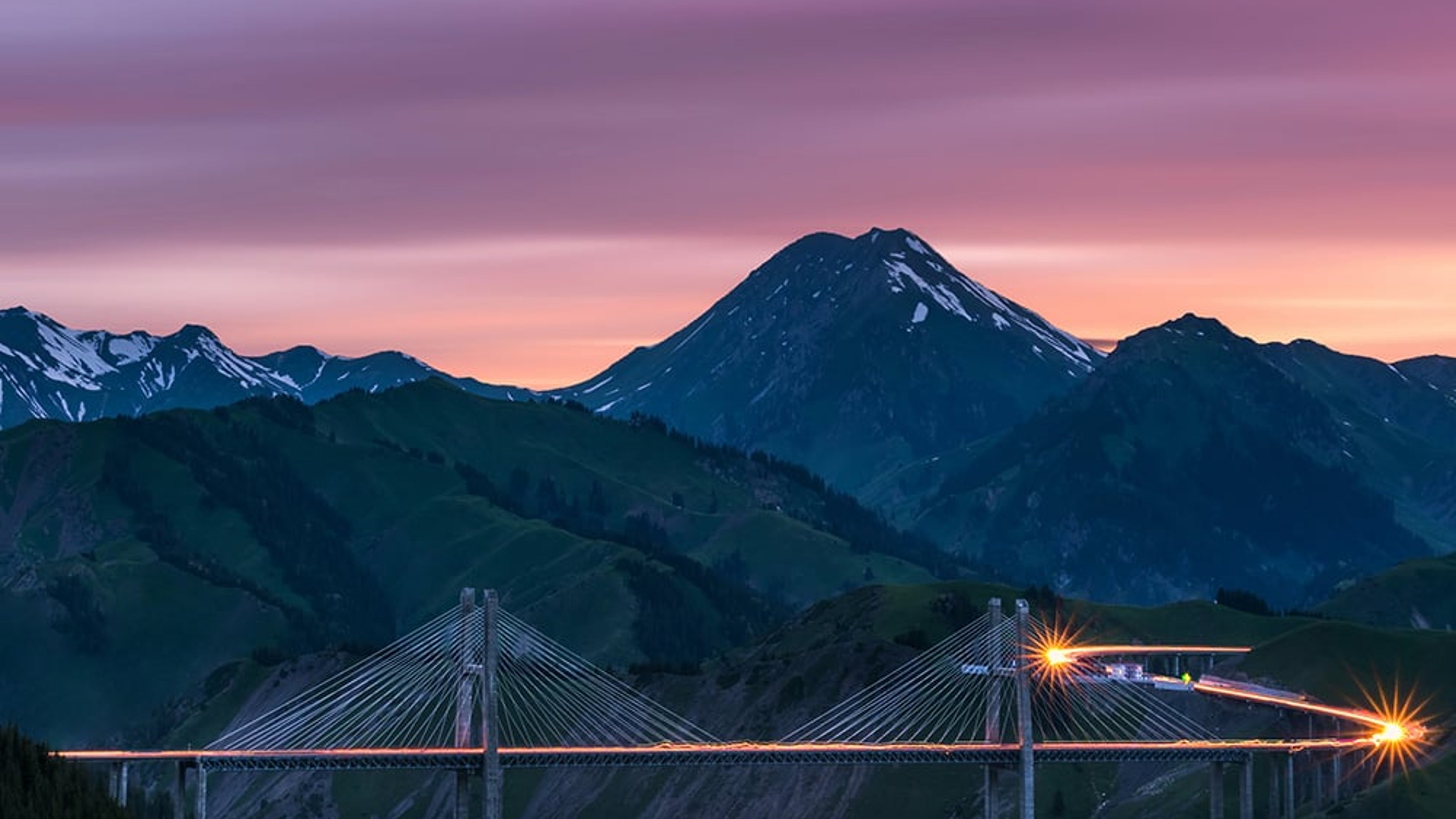 Xinjiang Guozigou Bridge in China with vehicle light trails and a colourful pink and purple sky at sunset or sunrise. Snow-capped peaks are visible in the distance.