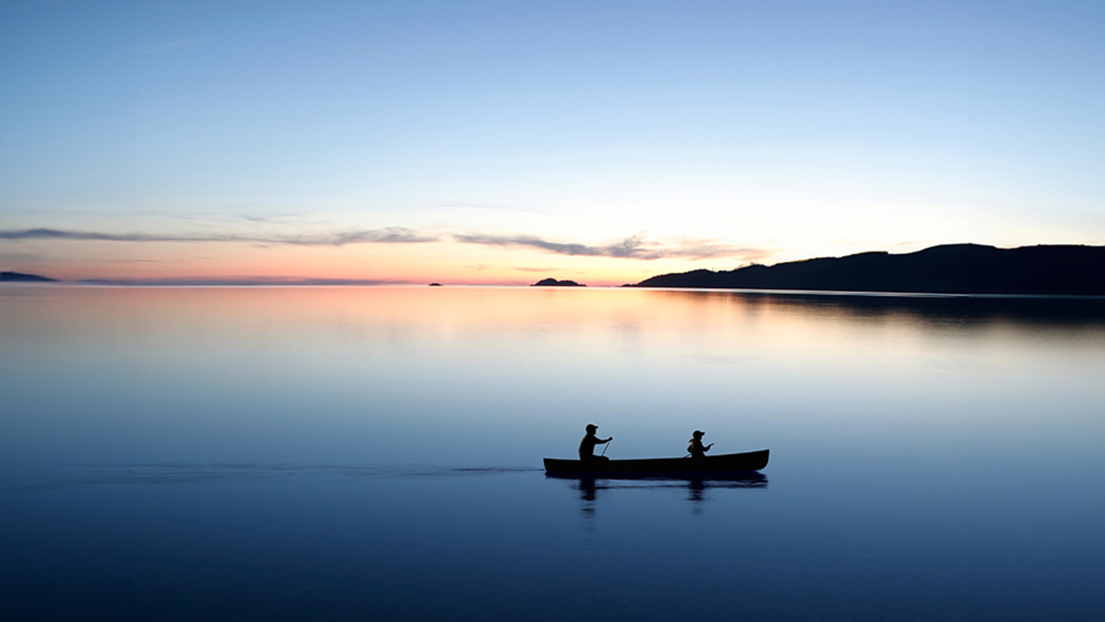 Lake Michigan at dusk, showing two people silhouetted in a canoe paddling across calm water, with a colourful sky and distant shoreline in the background.