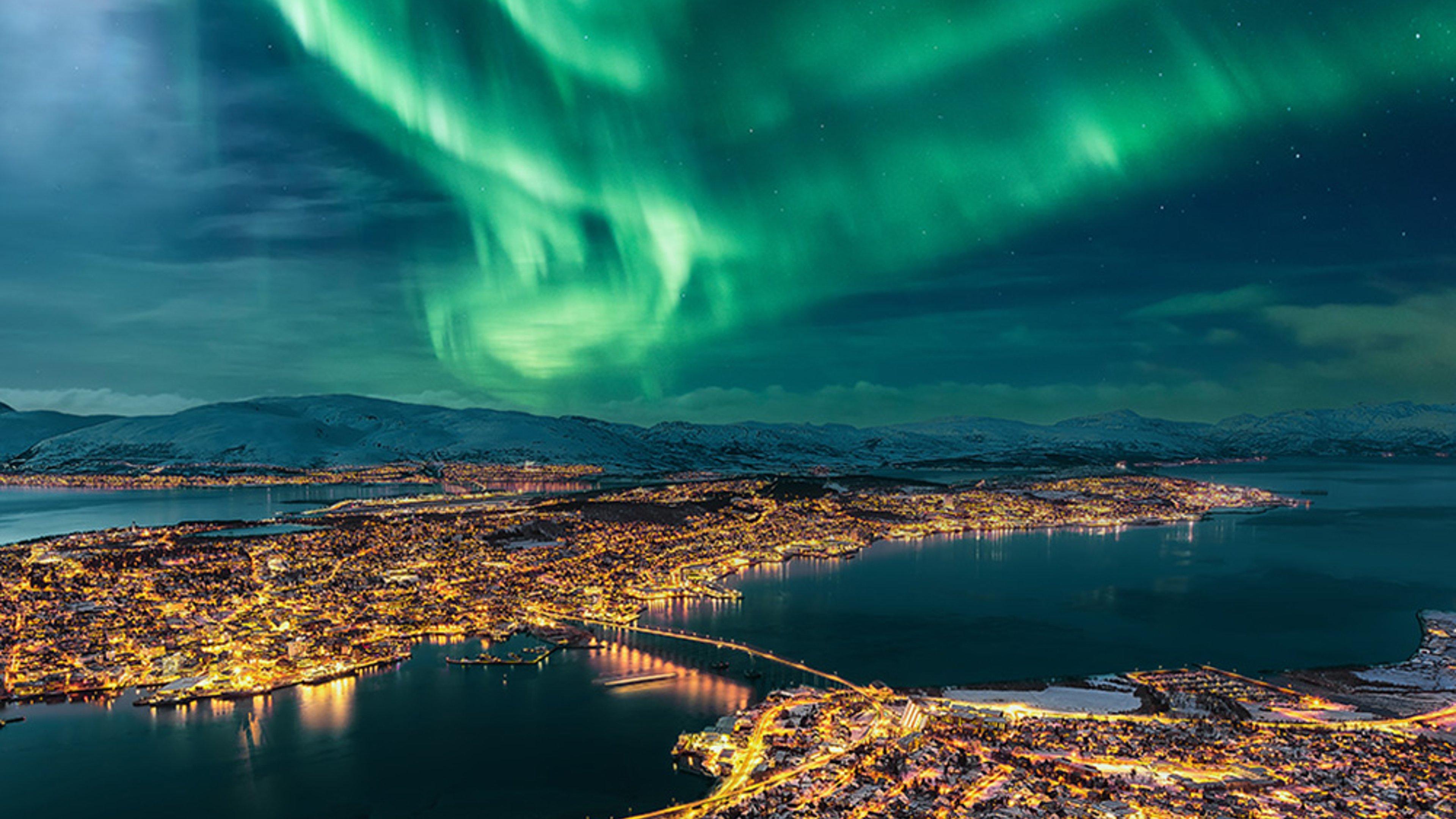 Aurora Borealis dancing in vibrant green hues over the city of Tromsø, Troms, northern Norway, with city lights reflecting on water and mountains faintly visible under the night sky.