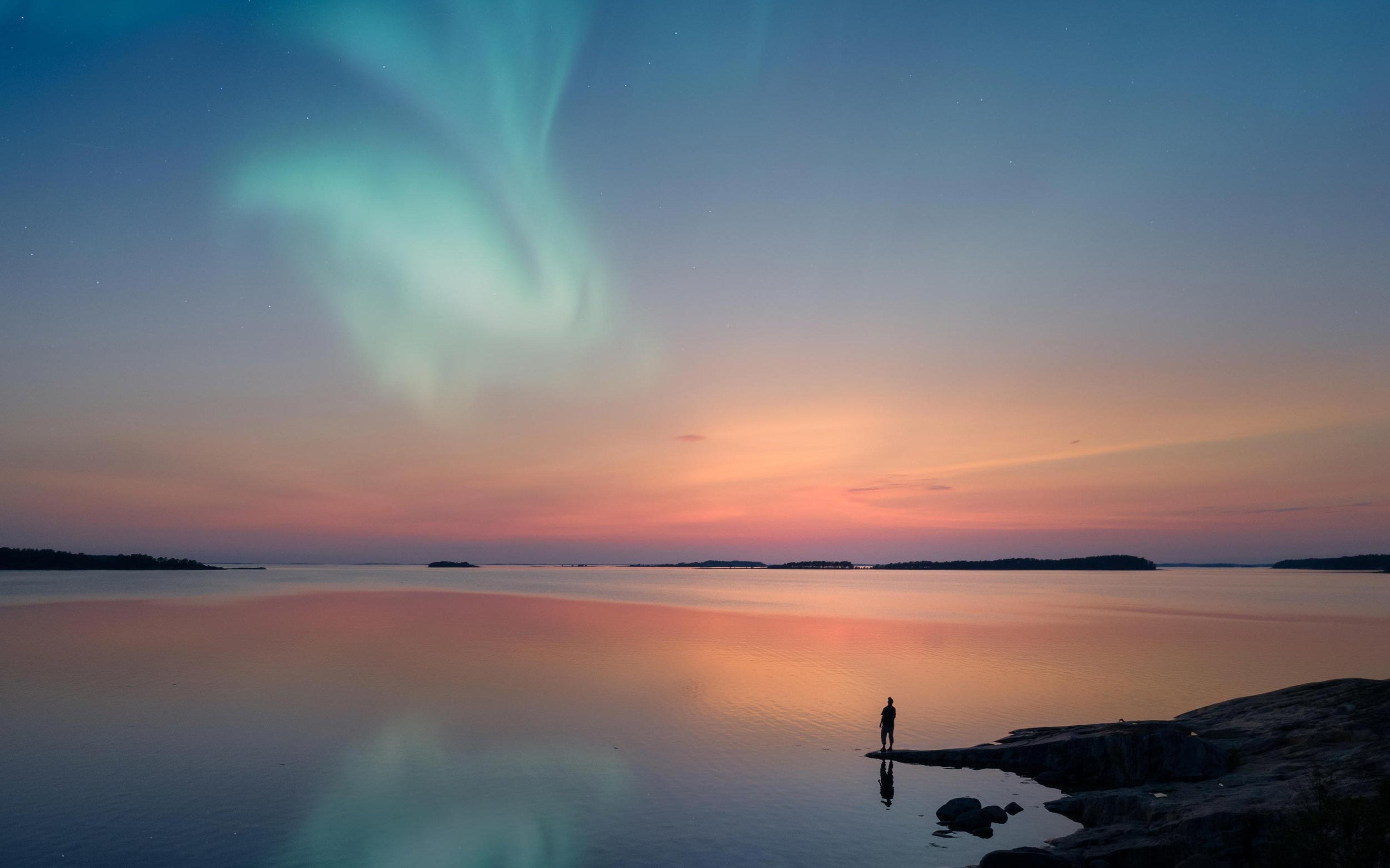 Silhouette of a man standing by a lake shore and looking at a beautiful aurora borealis on the sky with reflections on the calm lake.