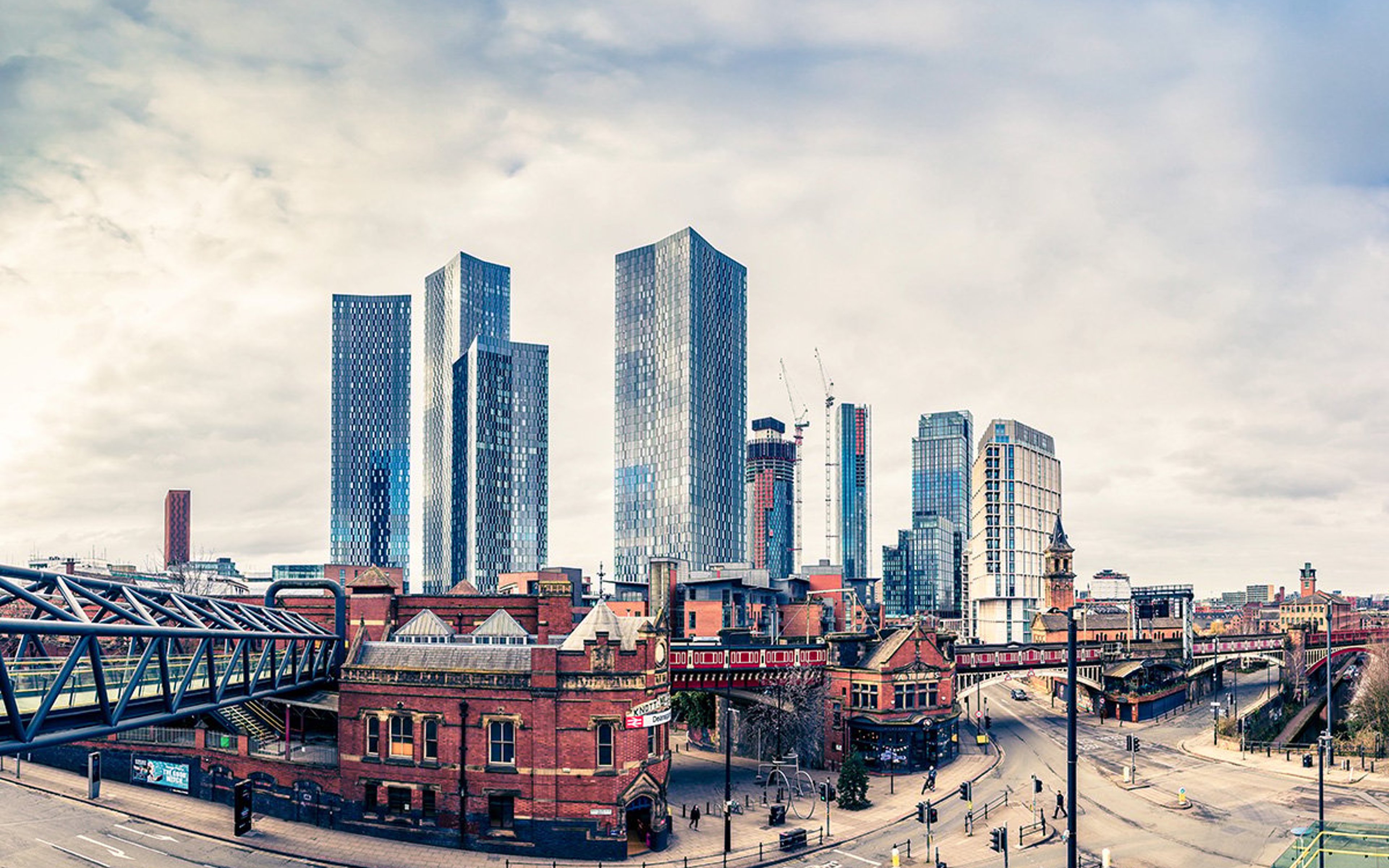 Panoramic view of Manchester city skyline in Lancashire, featuring tall glass skyscrapers under a cloudy sky, with historic red-brick buildings and a pedestrian bridge in the foreground.