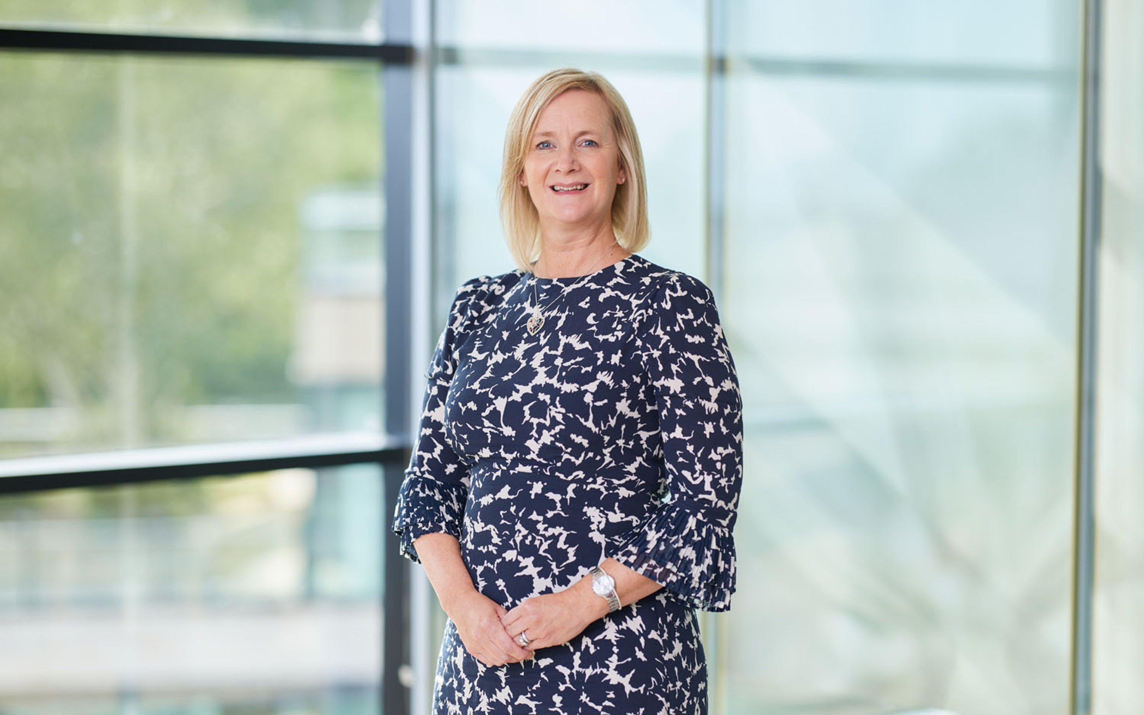 Angela Geddes wearing a blue floral print dress standing in front of an office window.