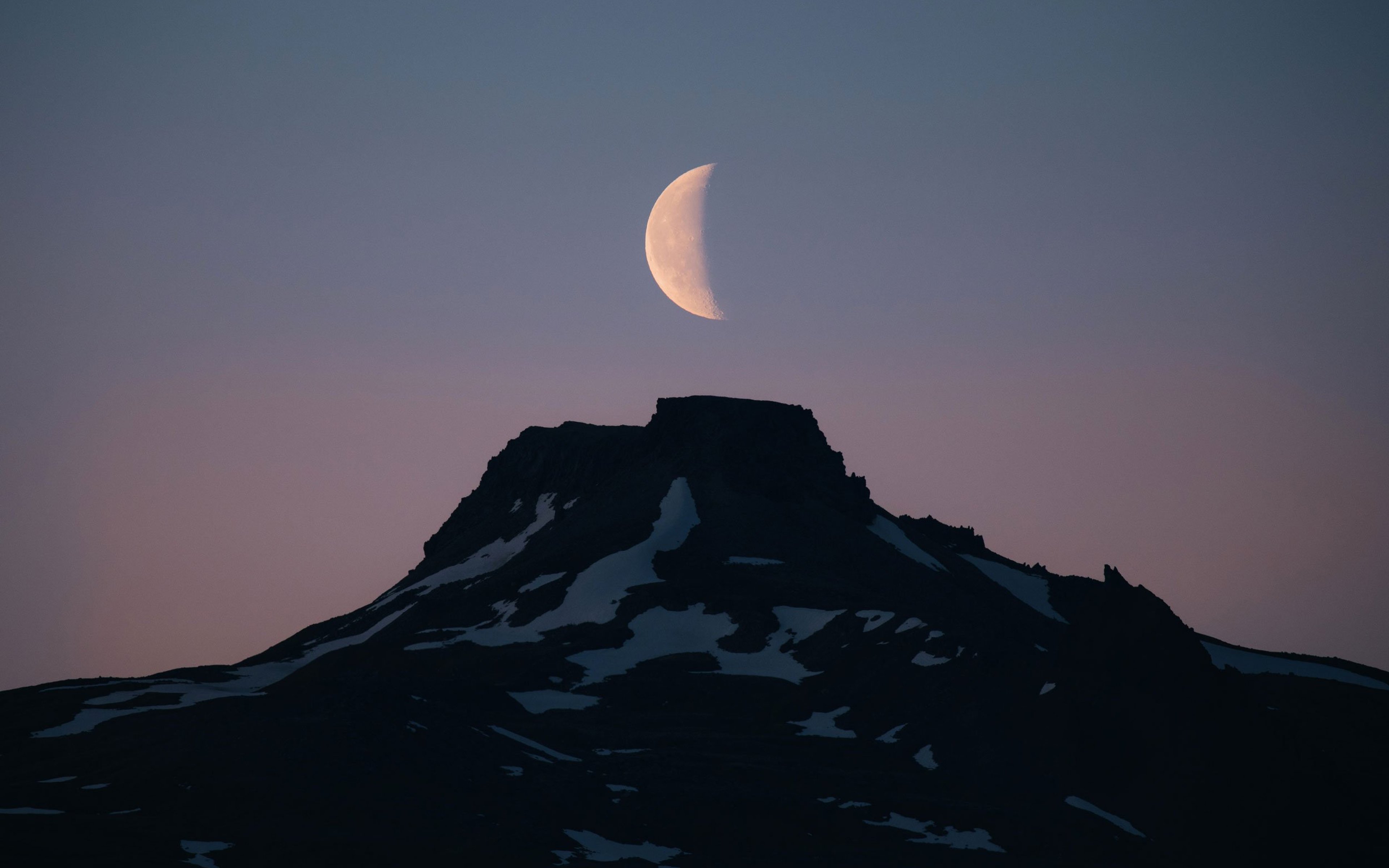 A half moon sits above a snowy mountain top at sunset.
