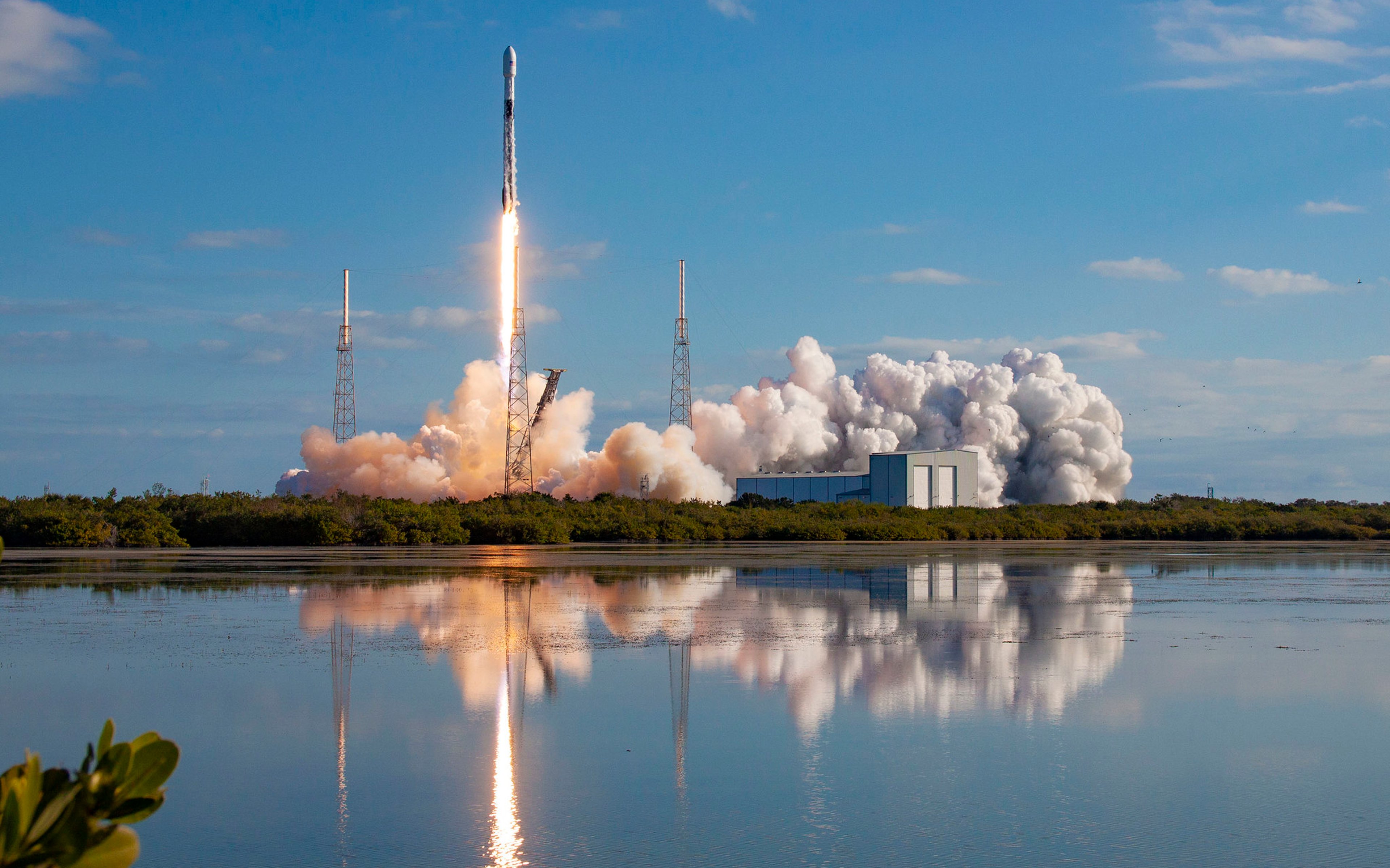 A SpaceX Falcon 9 rocket launches a Starlink mission from a coastal launchpad, with bright flames, thick smoke, and reflections visible in the water below.