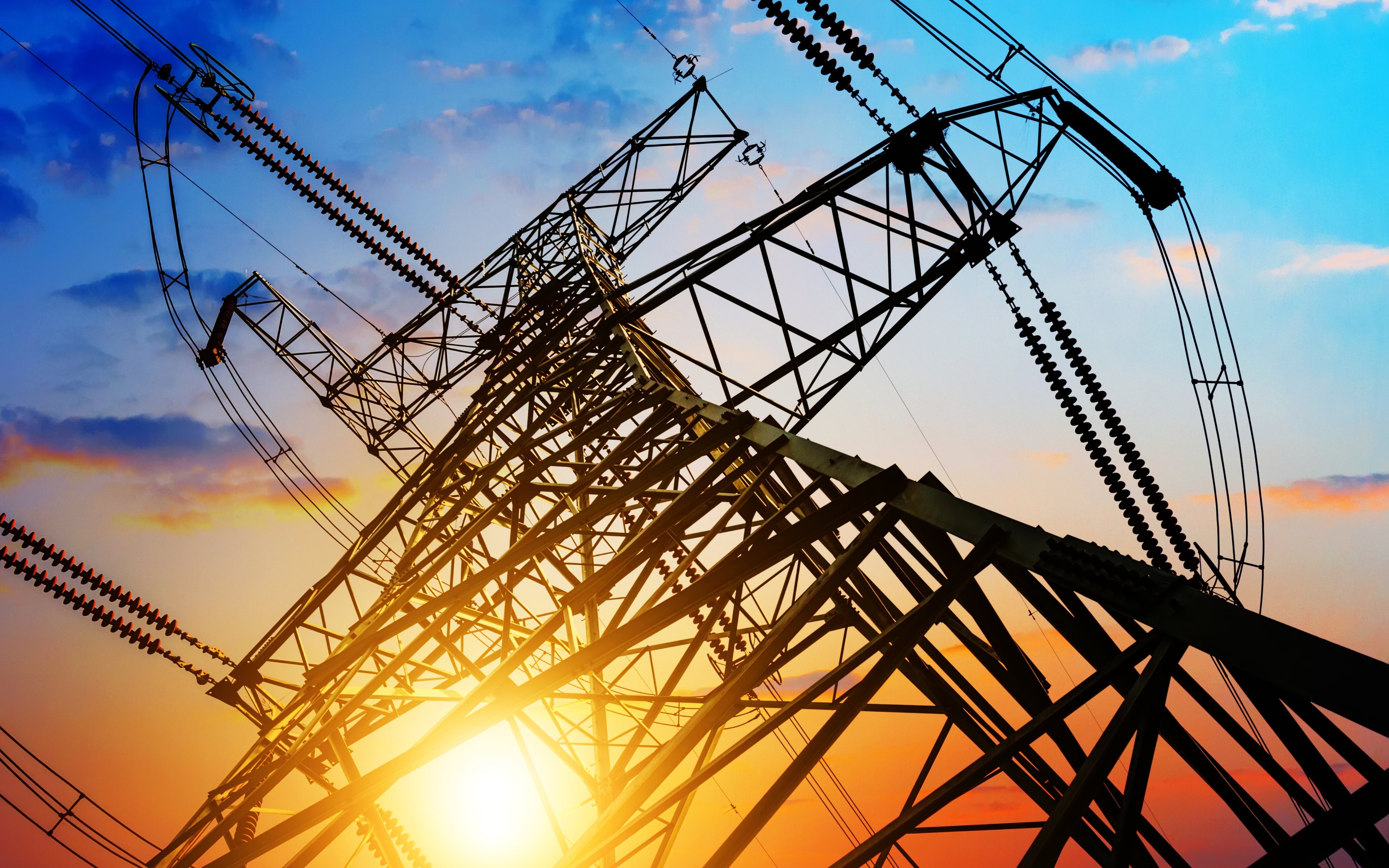 Low-angle view of a high-voltage electricity transmission tower silhouetted against a colourful sunset sky, with power lines and insulators stretching outward in multiple directions.