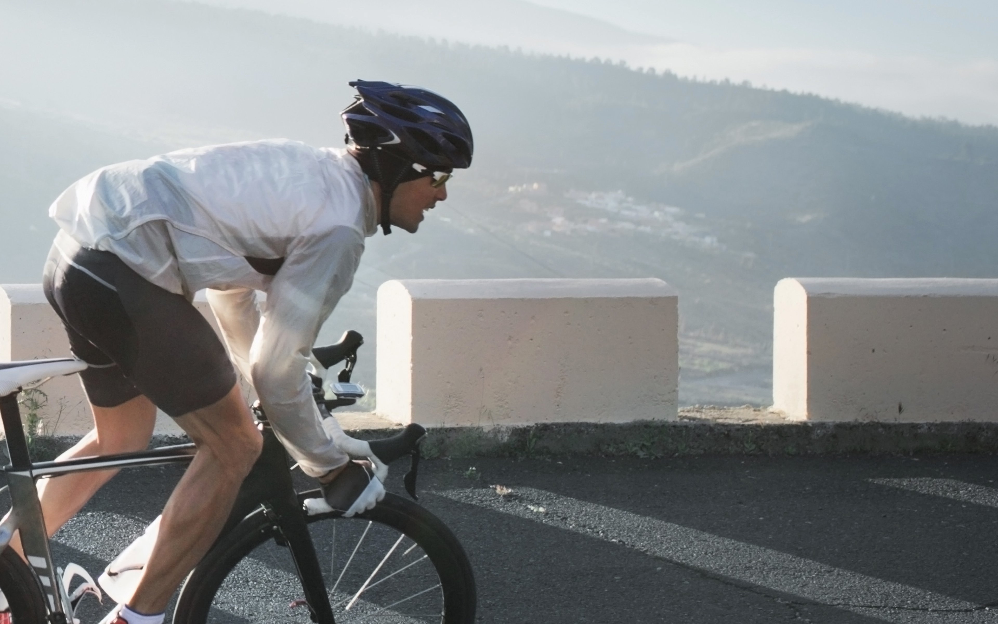 Photograph of a cyclist wearing a helmet and white jacket riding a road bike uphill on a mountain road with concrete barriers. Background shows misty hills and valleys, highlighting a challenging outdoor cycling activity.