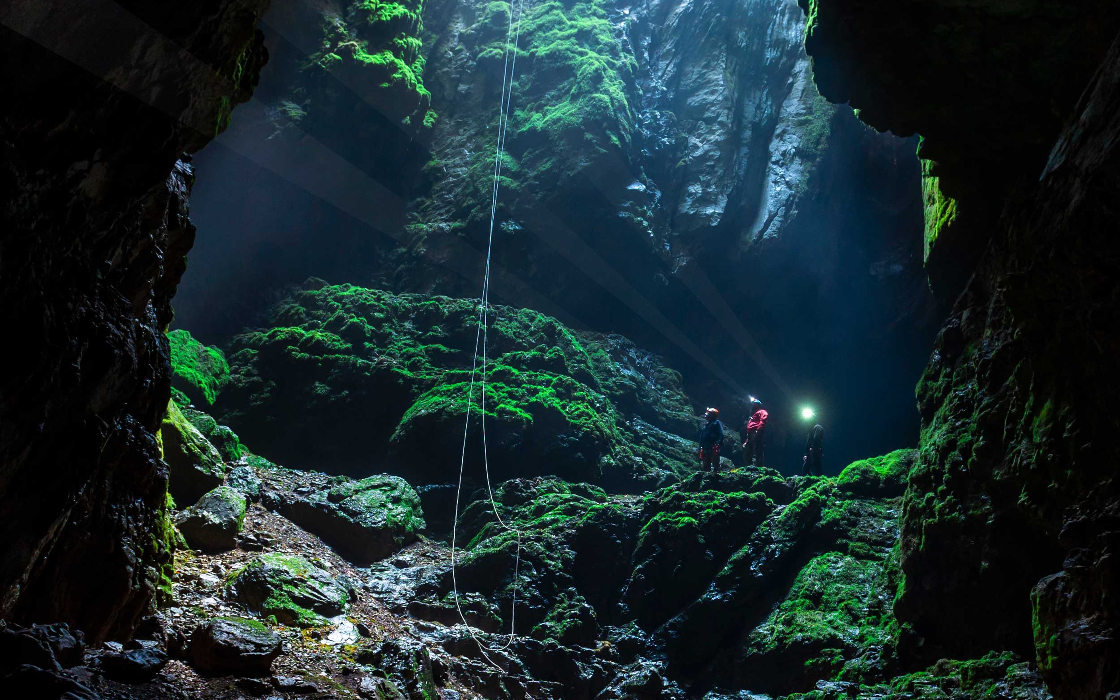 Caving team at the bottom of a deep cave wearing torches, with ropes hanging from the daylight above.