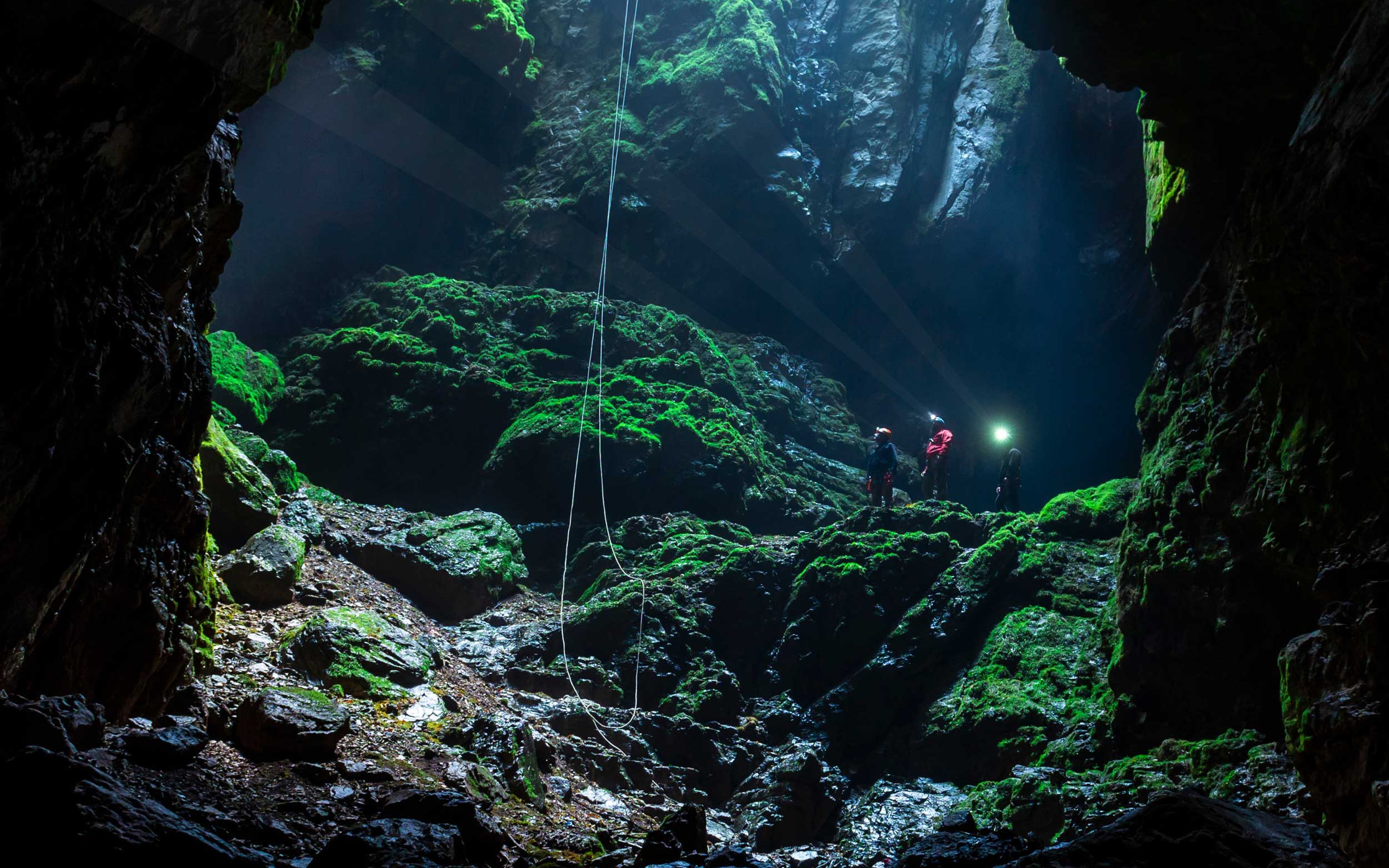 Caving team at the bottom of a deep cave wearing torches, with ropes hanging from the daylight above.
