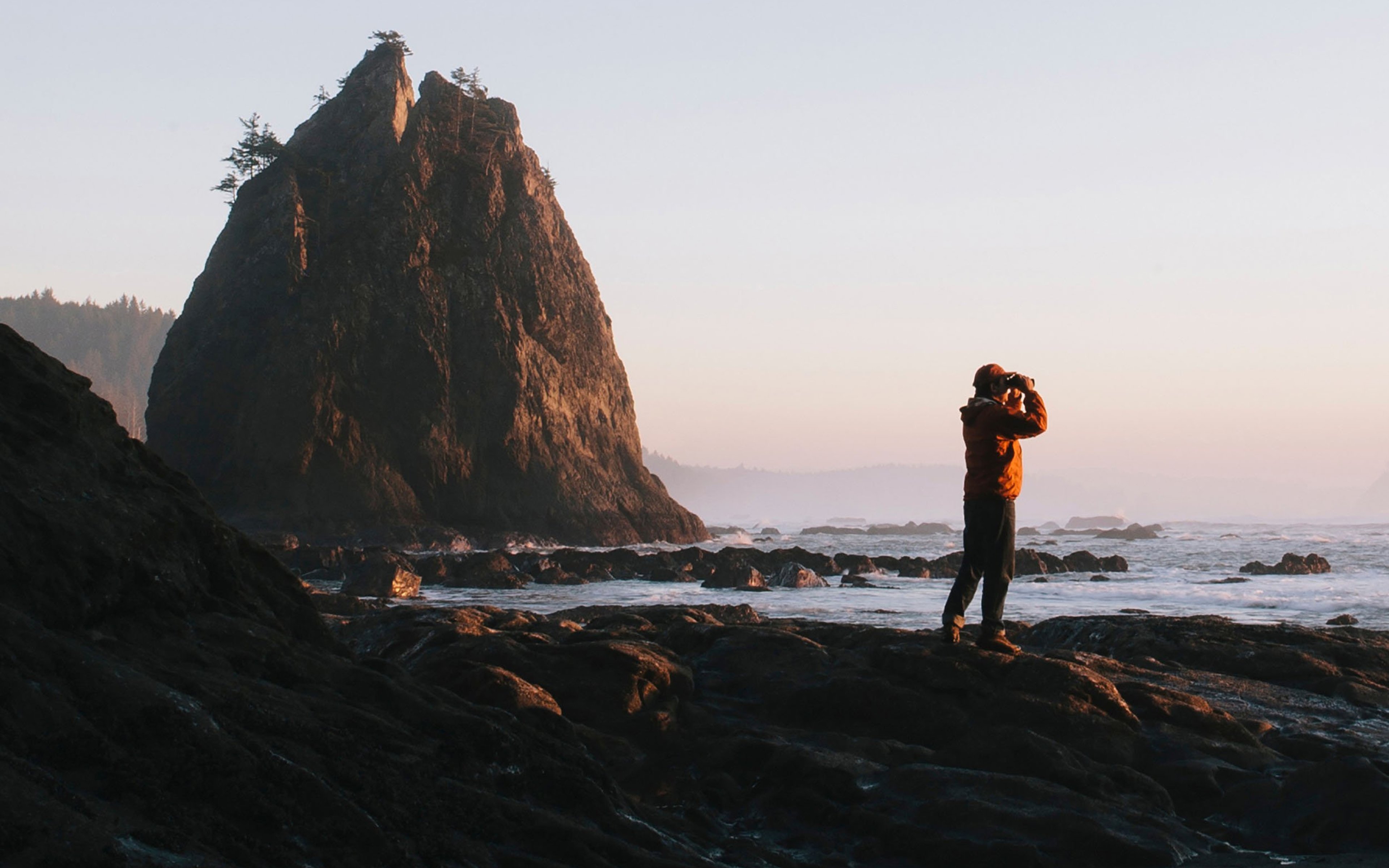 Man standing beneath sea cliff at dusk, using binoculars.