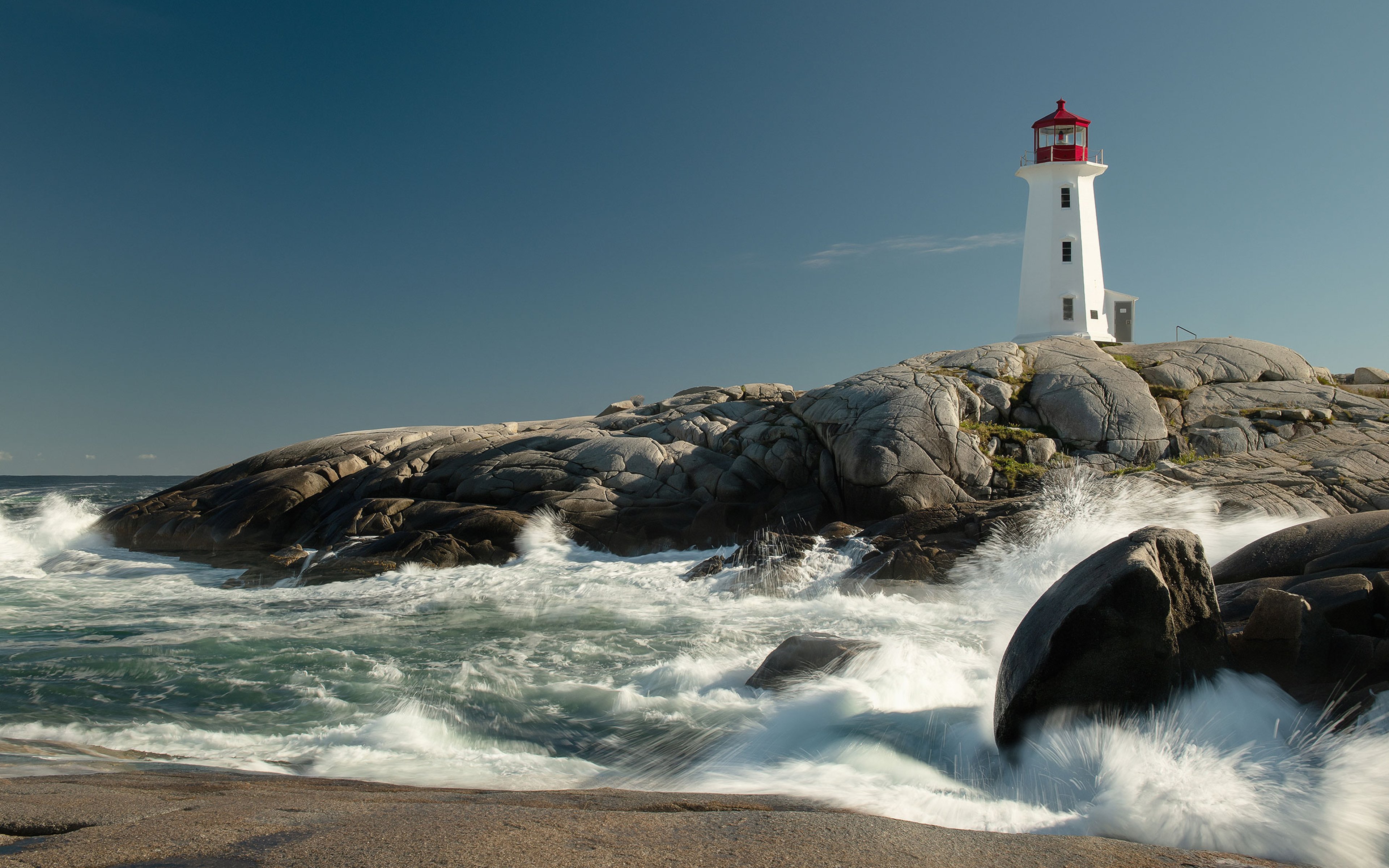 A lighthouse on top of rocks on a clear day with waves crashing in the foreground.