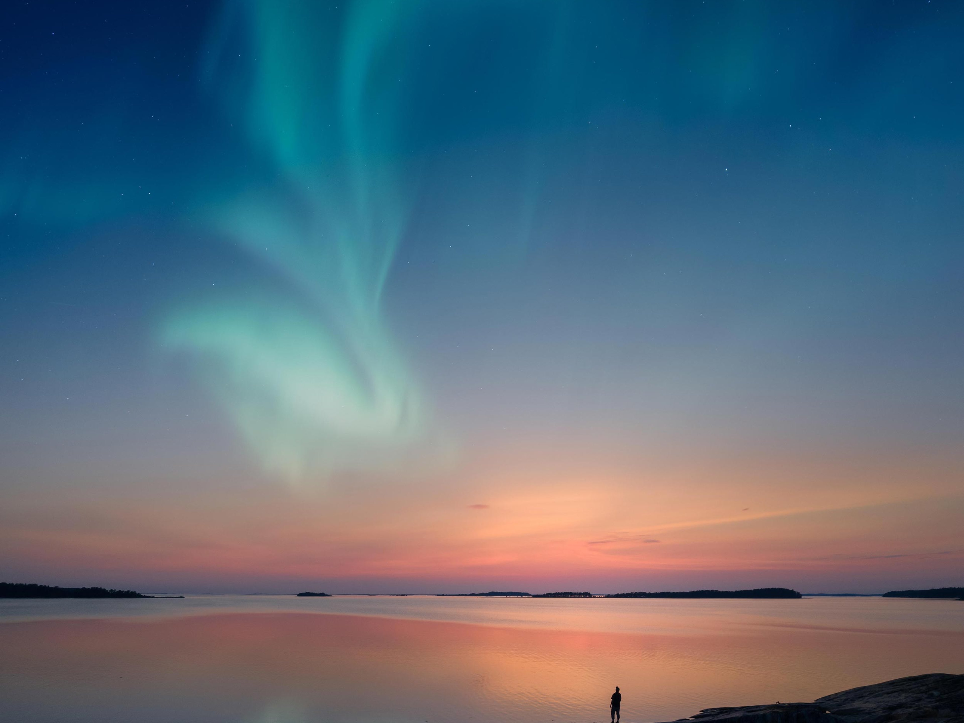 Silhouette of a man standing by a lake shore and looking at a beautiful aurora borealis on the sky with reflections on the calm lake.