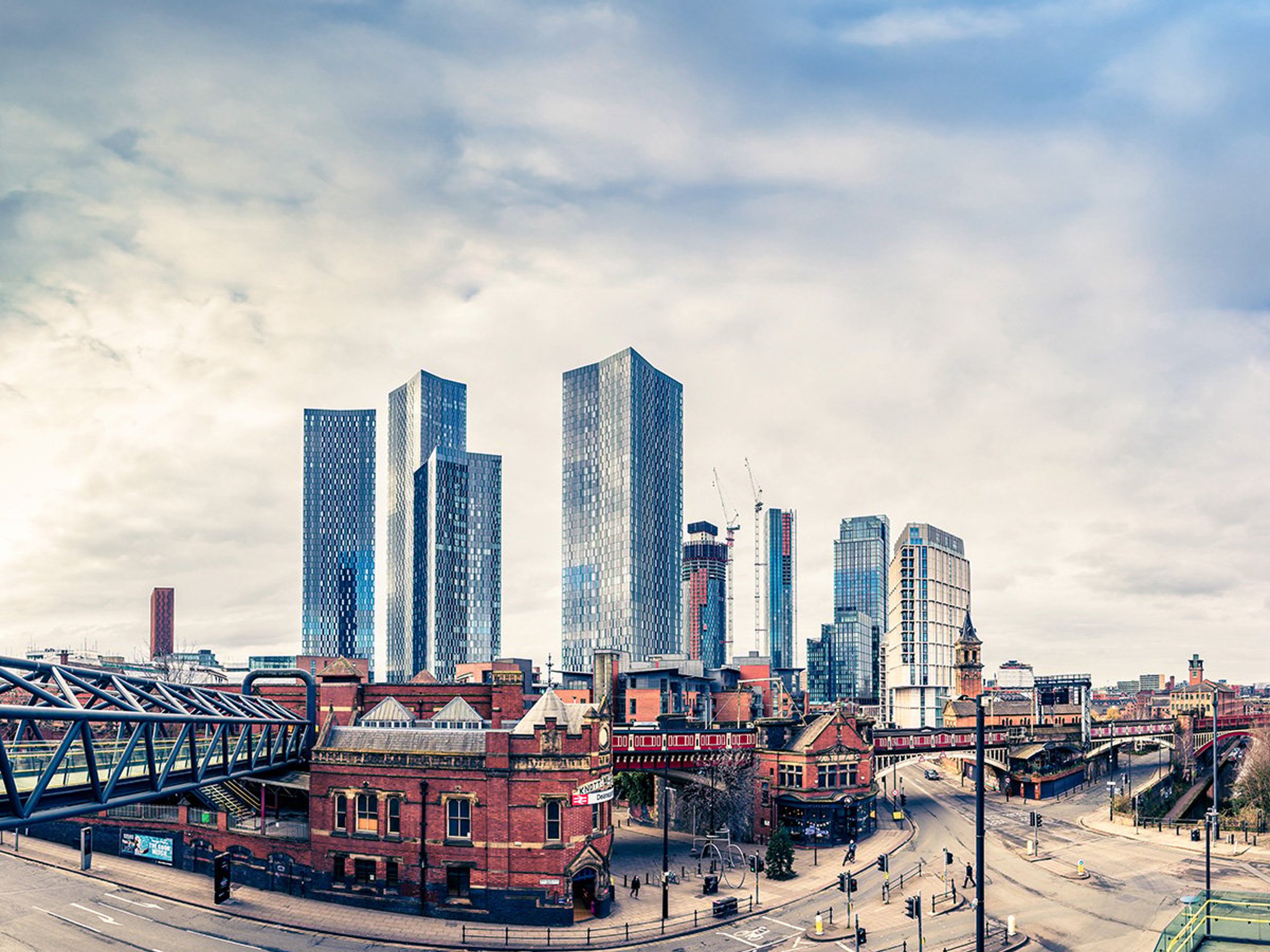 Panoramic view of Manchester city skyline in Lancashire, featuring tall glass skyscrapers under a cloudy sky, with historic red-brick buildings and a pedestrian bridge in the foreground.