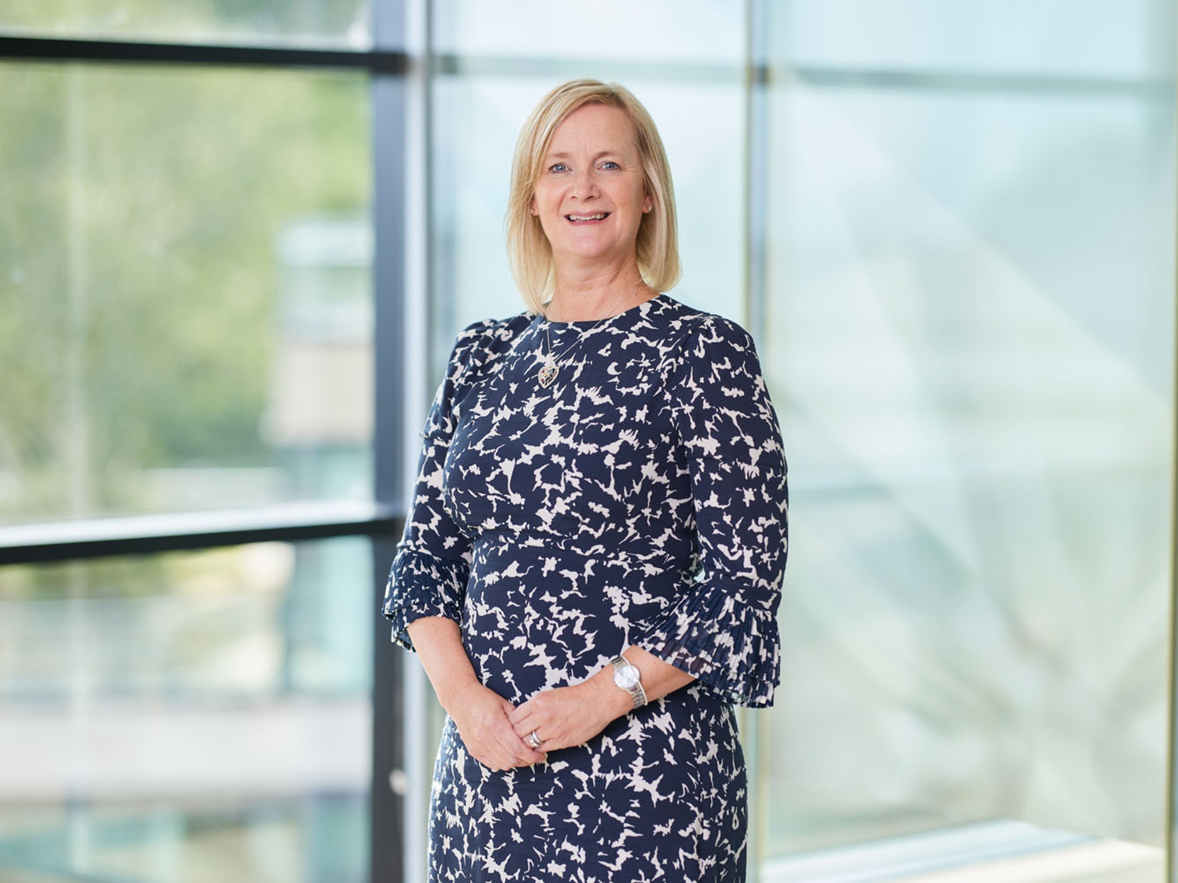 Angela Geddes wearing a blue floral print dress standing in front of an office window.
