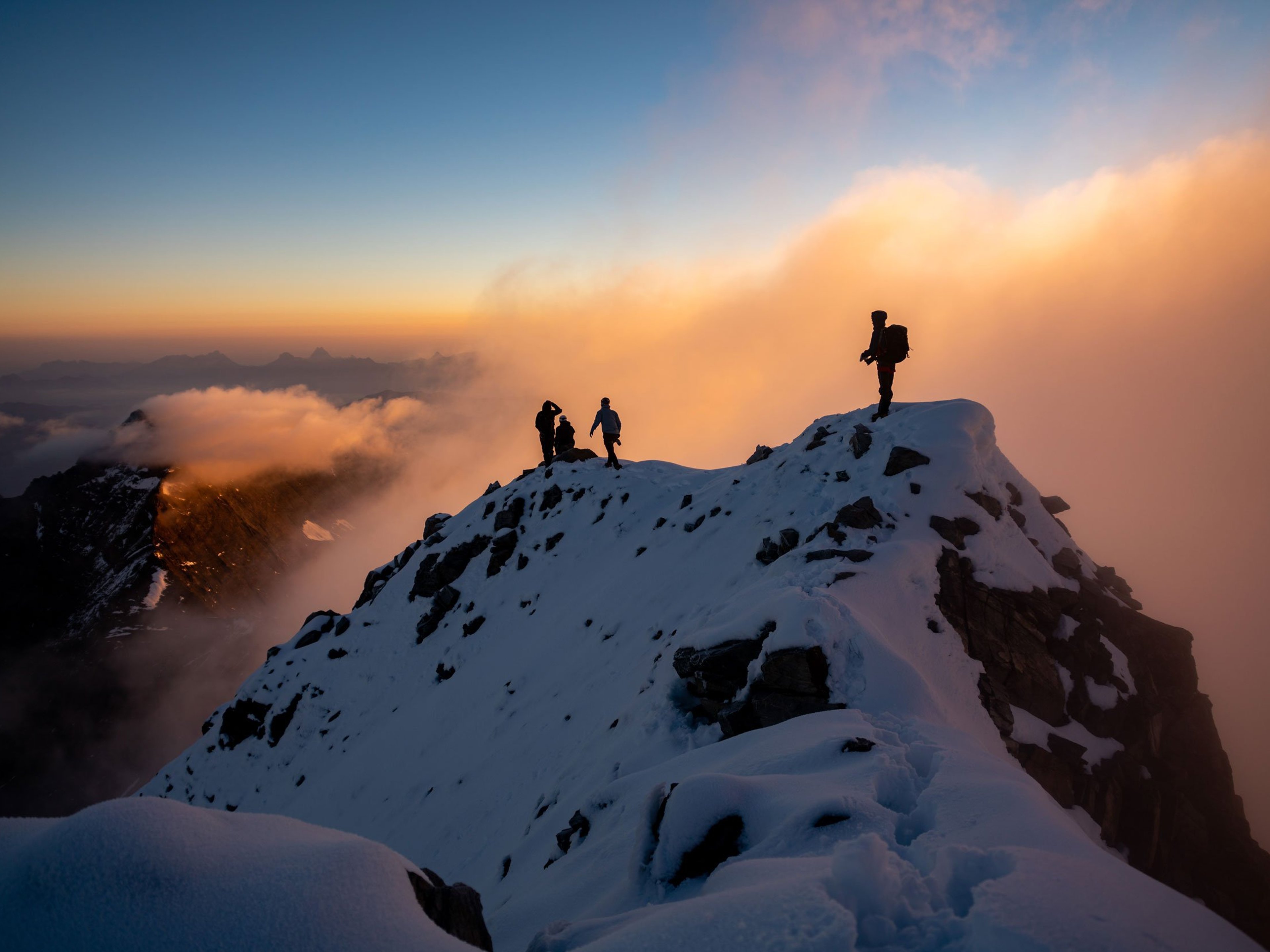 The sillouettes of four climbers at the summit of a snowy mountaintop at sunset.