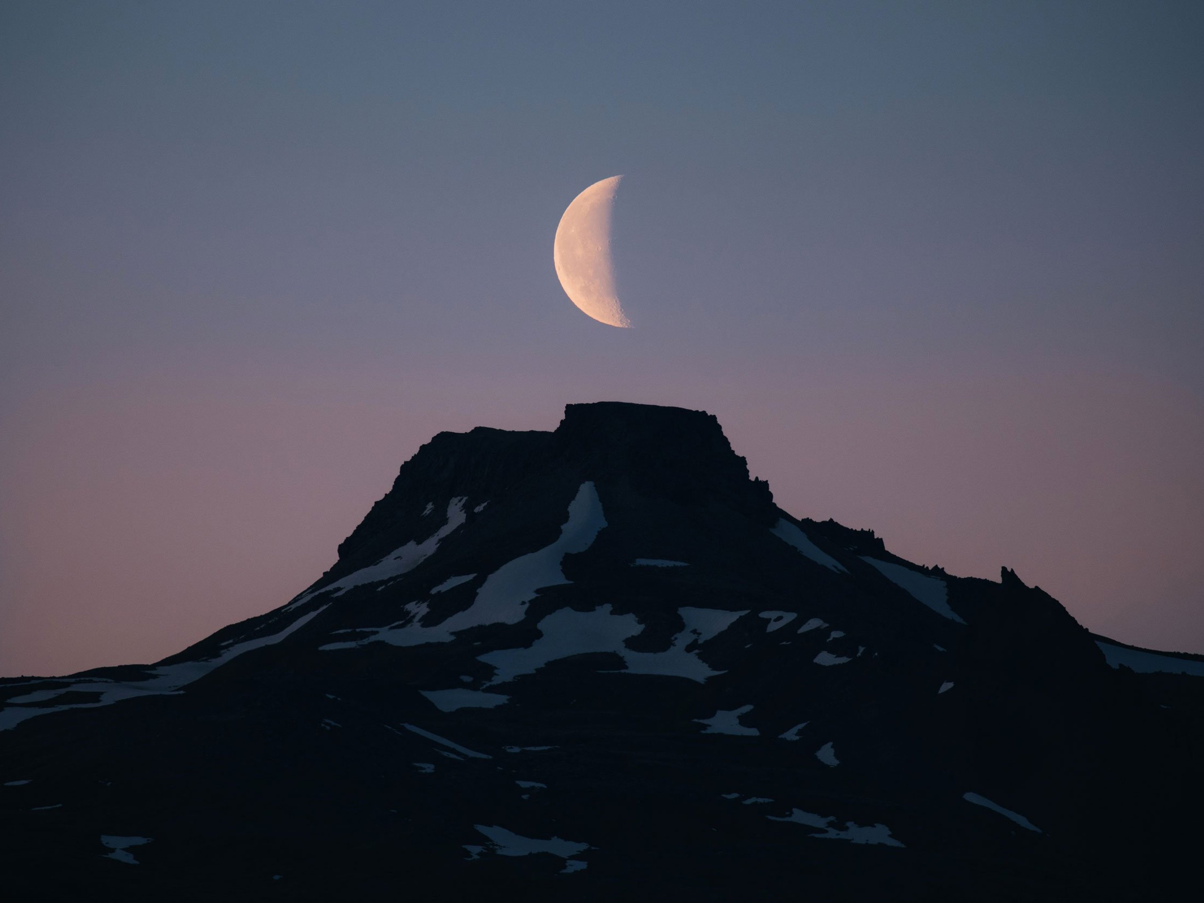 A half moon sits above a snowy mountain top at sunset.