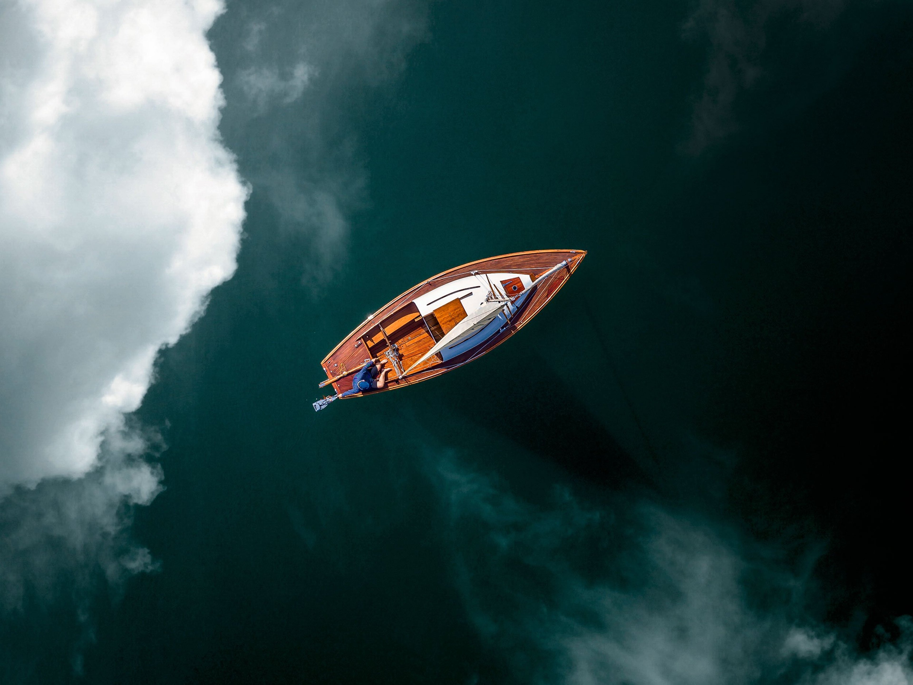 An aerial view of a sailing boat floating on still water with clouds reflected on the surface.