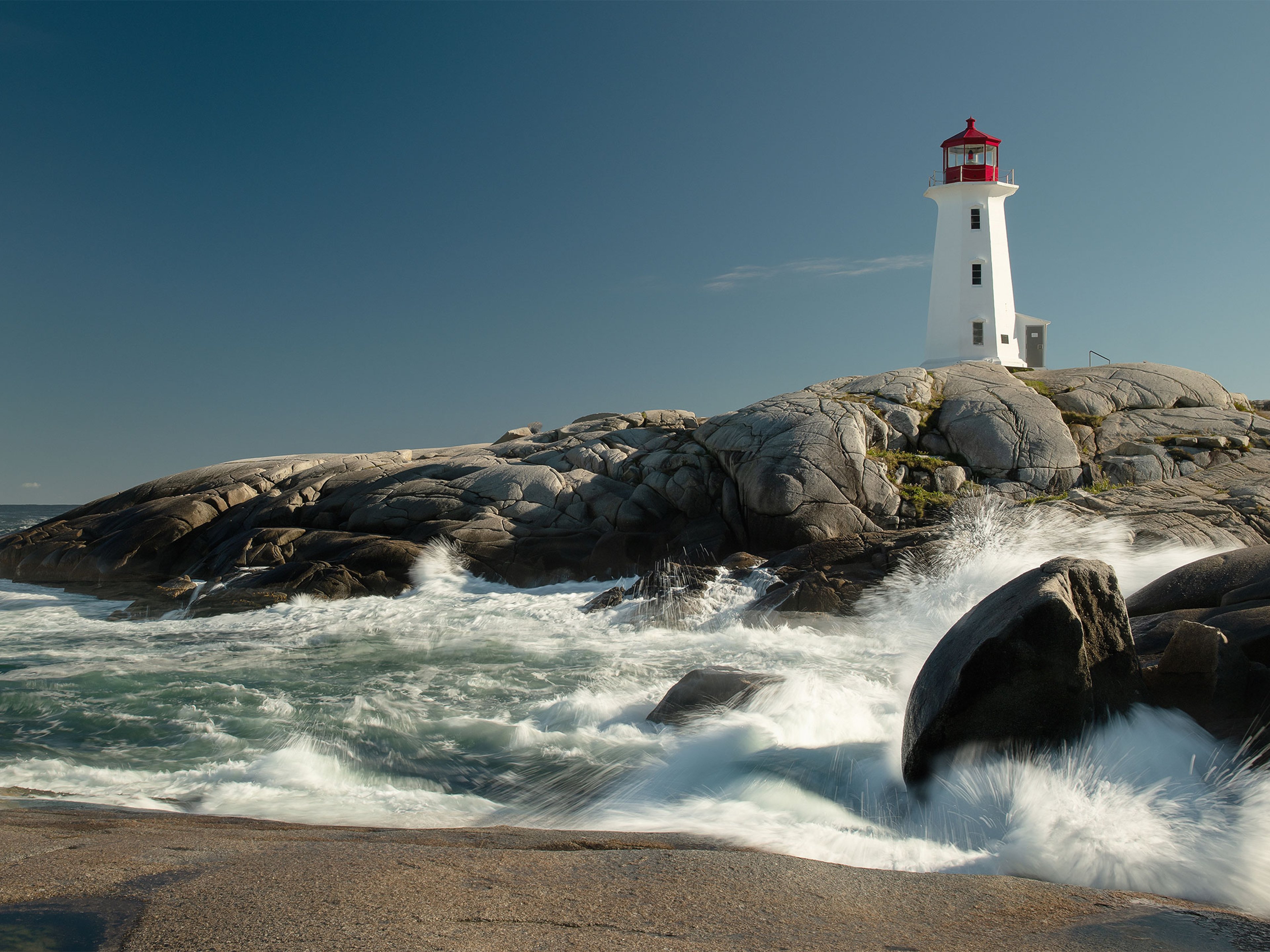 A lighthouse on top of rocks on a clear day with waves crashing in the foreground.