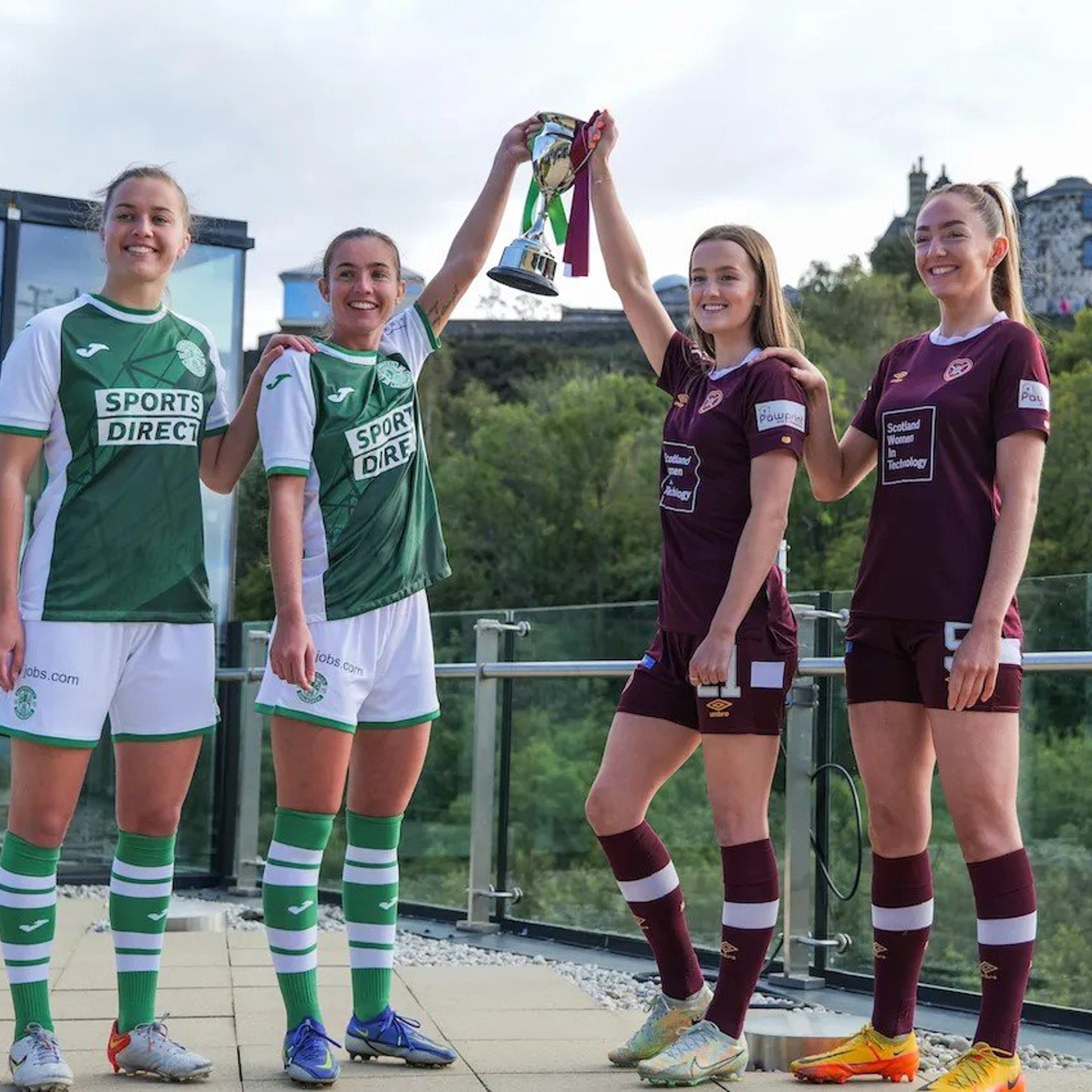 Two players from each of the Hibernian and Hearts woman's football teams lift a trophy while standing on a rooftop terrace.