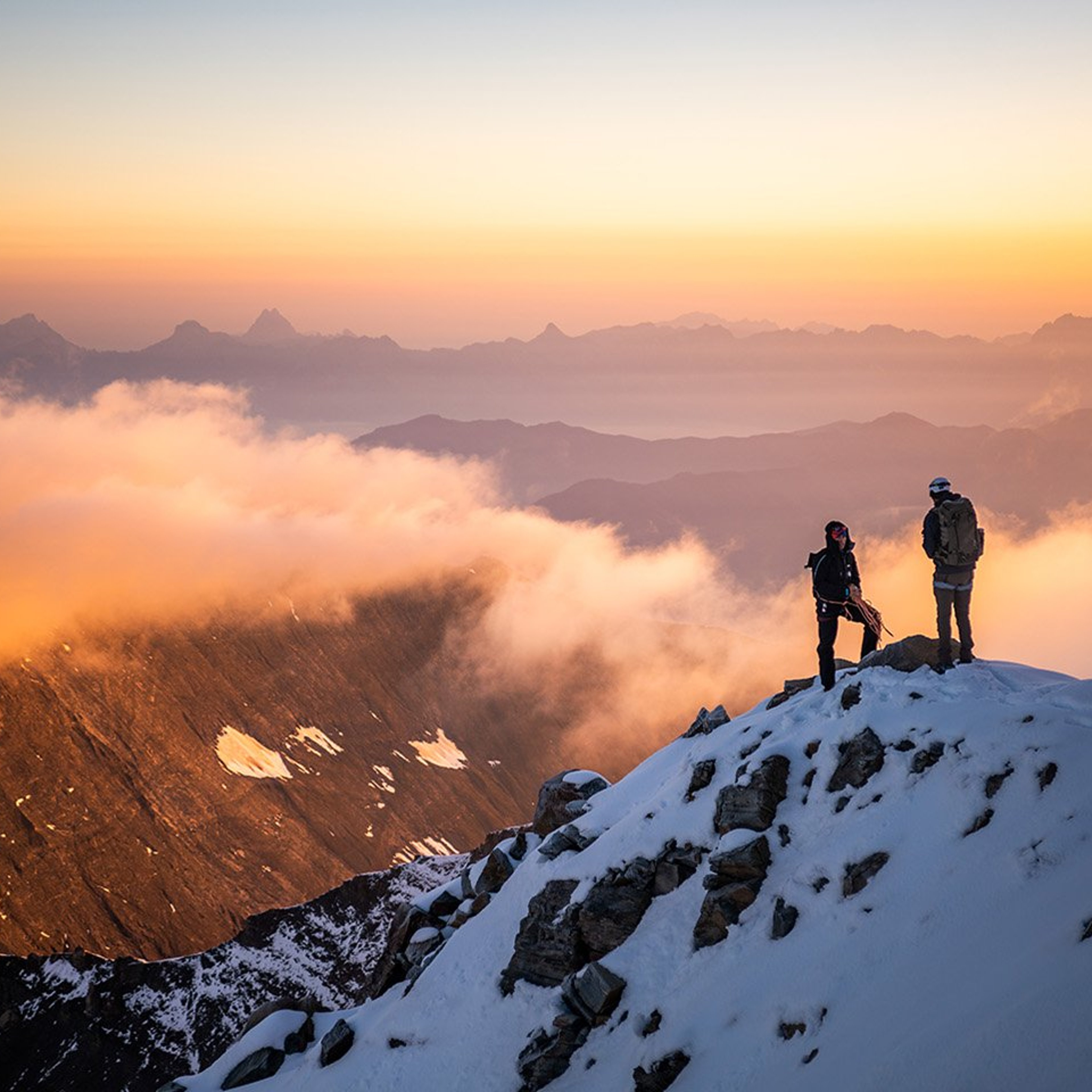 Two figures stand on the peak on a snow coverd mountain against a dramtic sunrise sky