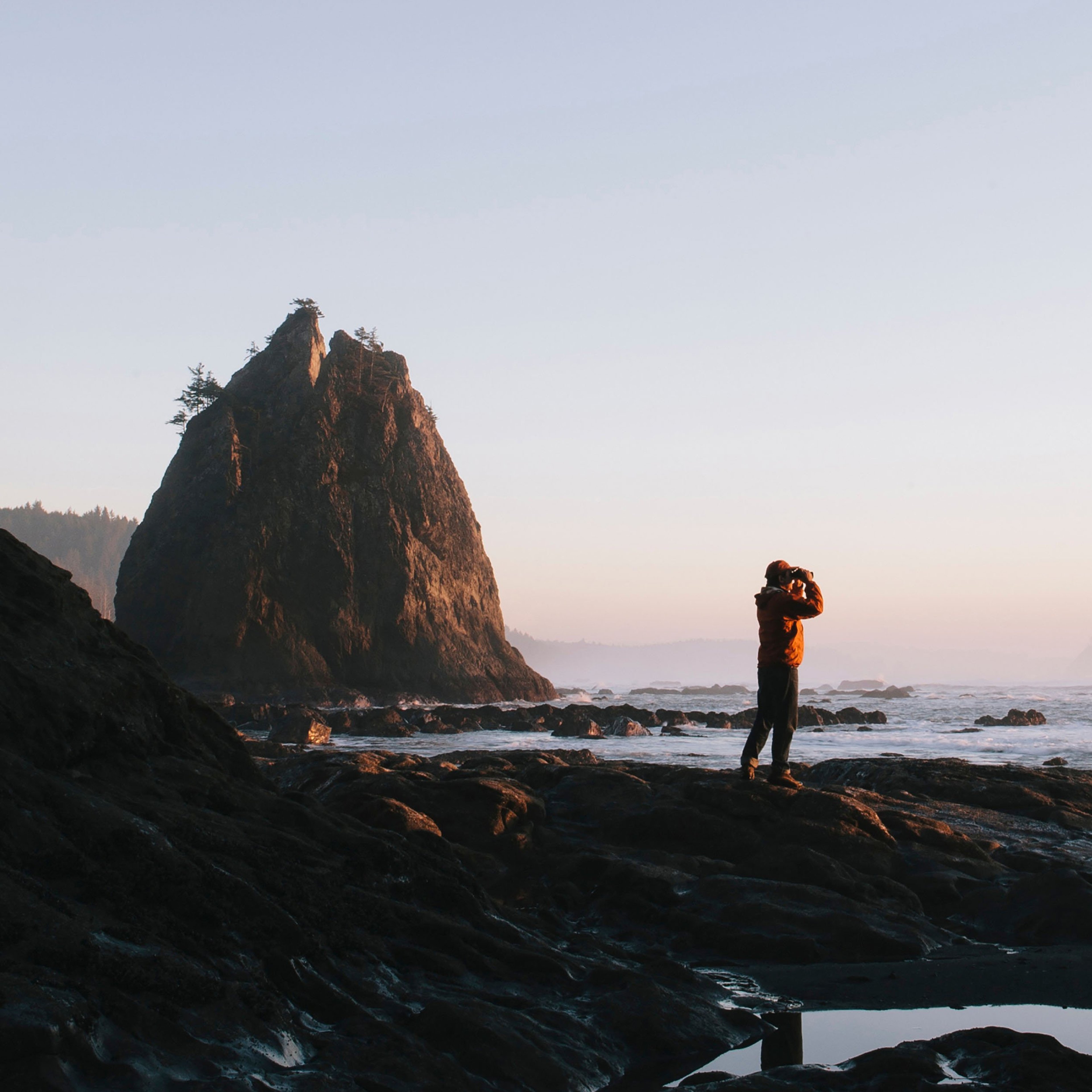 Man standing beneath sea cliff at dusk, using binoculars.